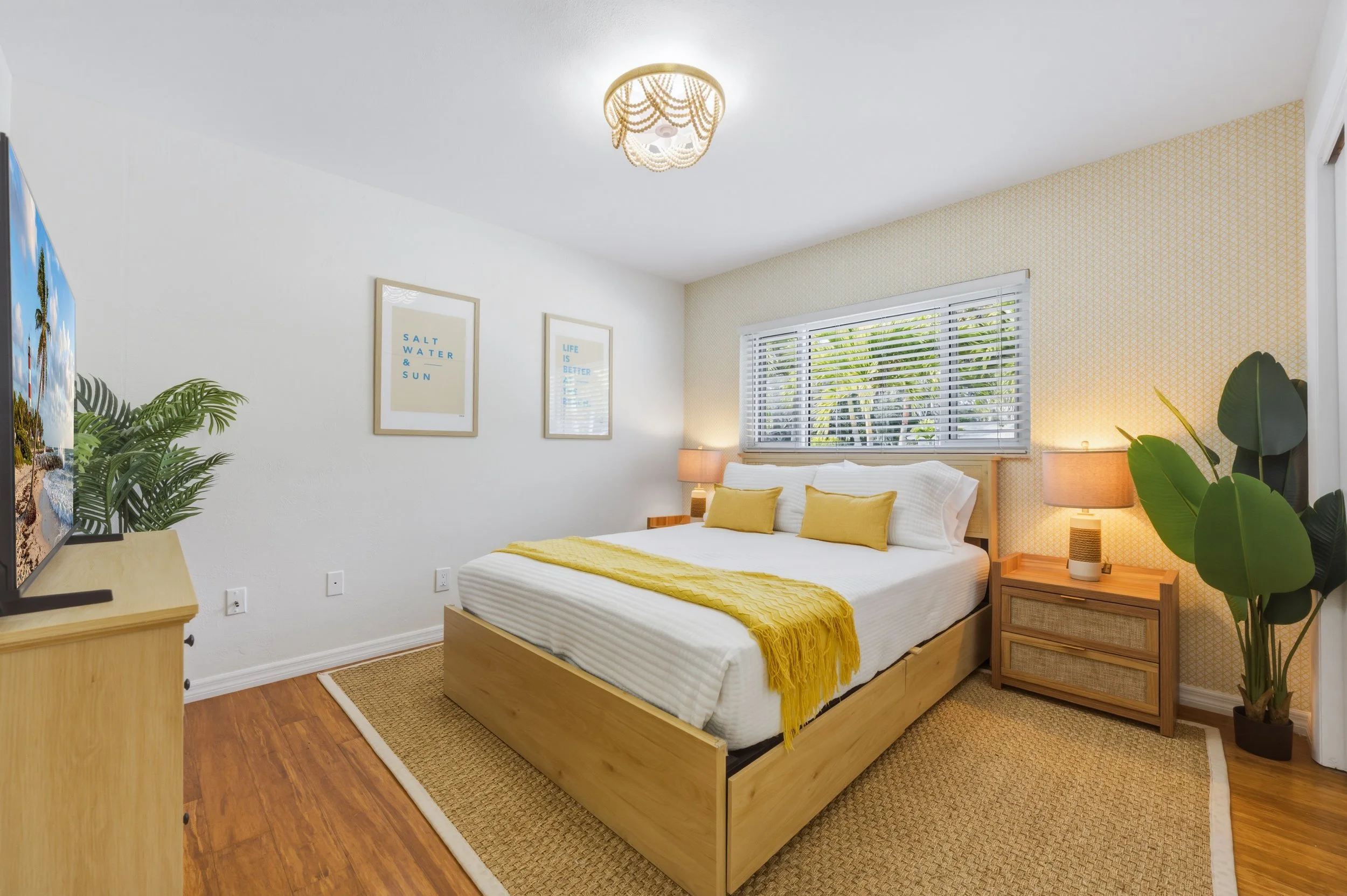 A cozy bedroom with a large bed, yellow pillows, and a yellow throw blanket. The room has wooden furniture, a window with blinds, a ceiling light fixture, and two bedside lamps. Decor includes framed art on the white wall and large green plants.