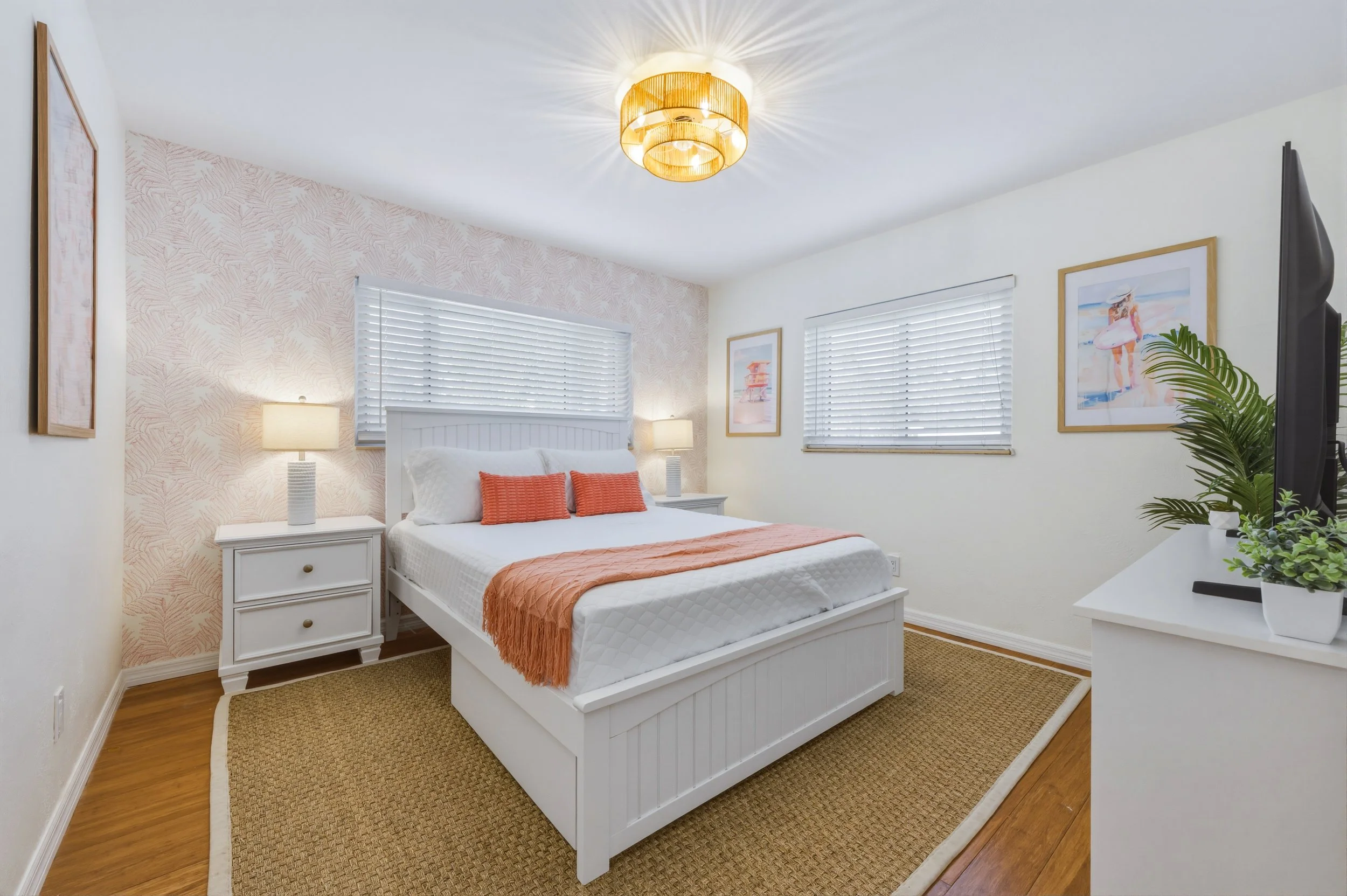 A cozy bedroom with a white bed, orange accents, two nightstands with lamps, framed artwork on the walls, and a flatscreen TV on a white dresser, with a ceiling light fixture.