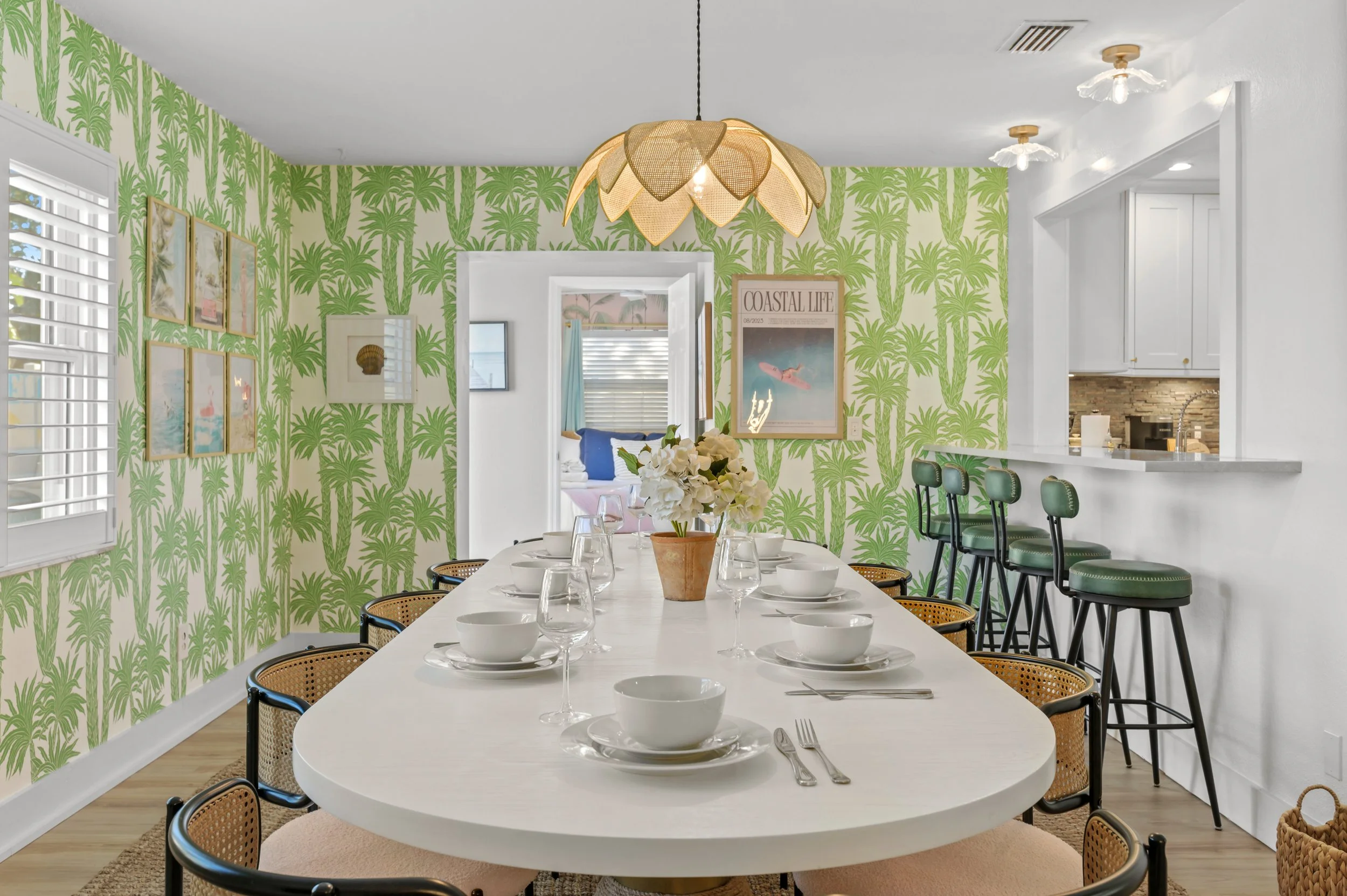 Dining room with a large white table set for a meal, green palm leaf patterned wallpaper, framed artwork, a flower centerpiece, and a bar area with green bar stools.