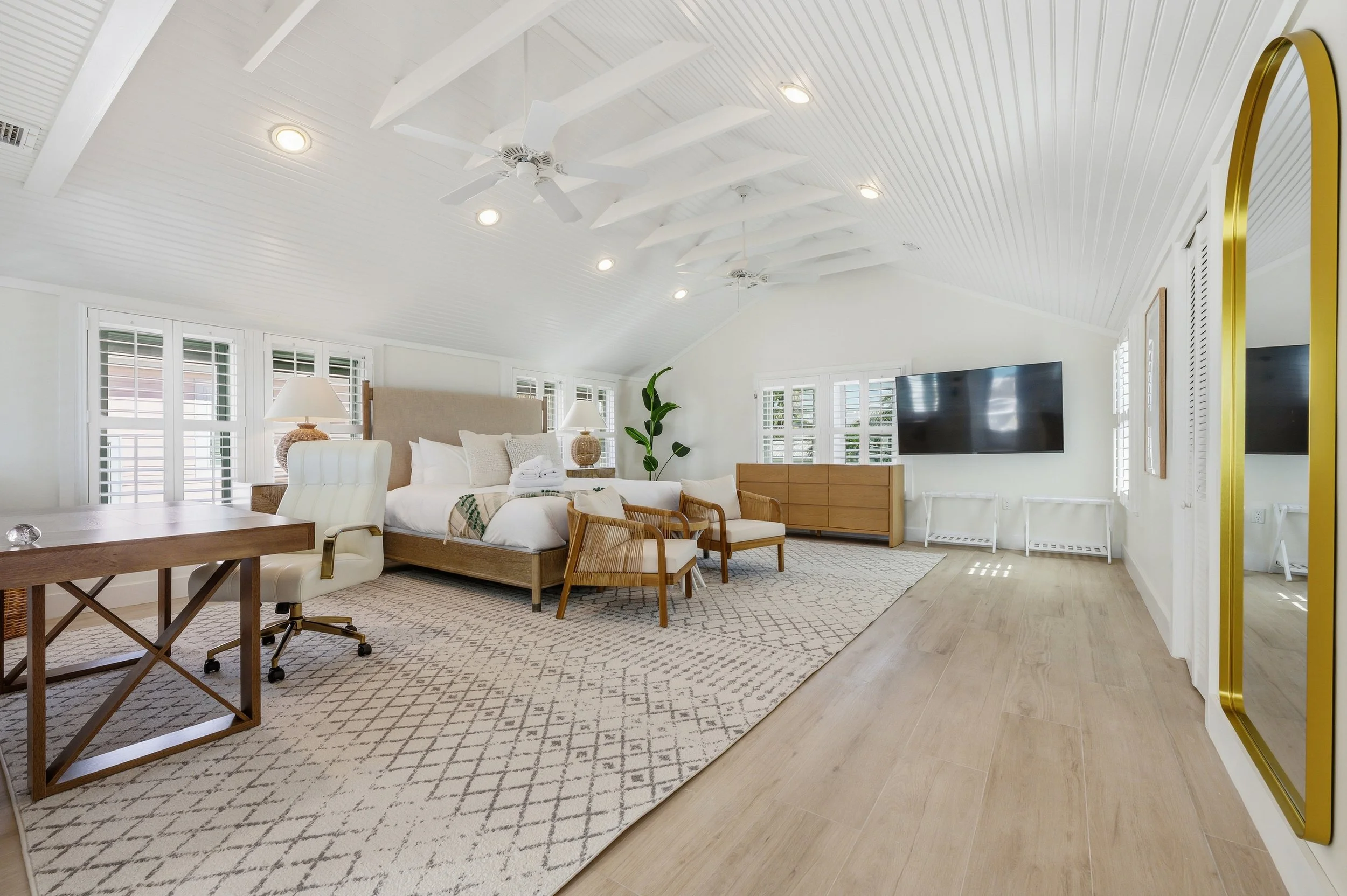 Spacious bedroom with white walls and ceiling, featuring a large bed with white linens, wooden furniture, a flat-screen TV, and a patterned rug.