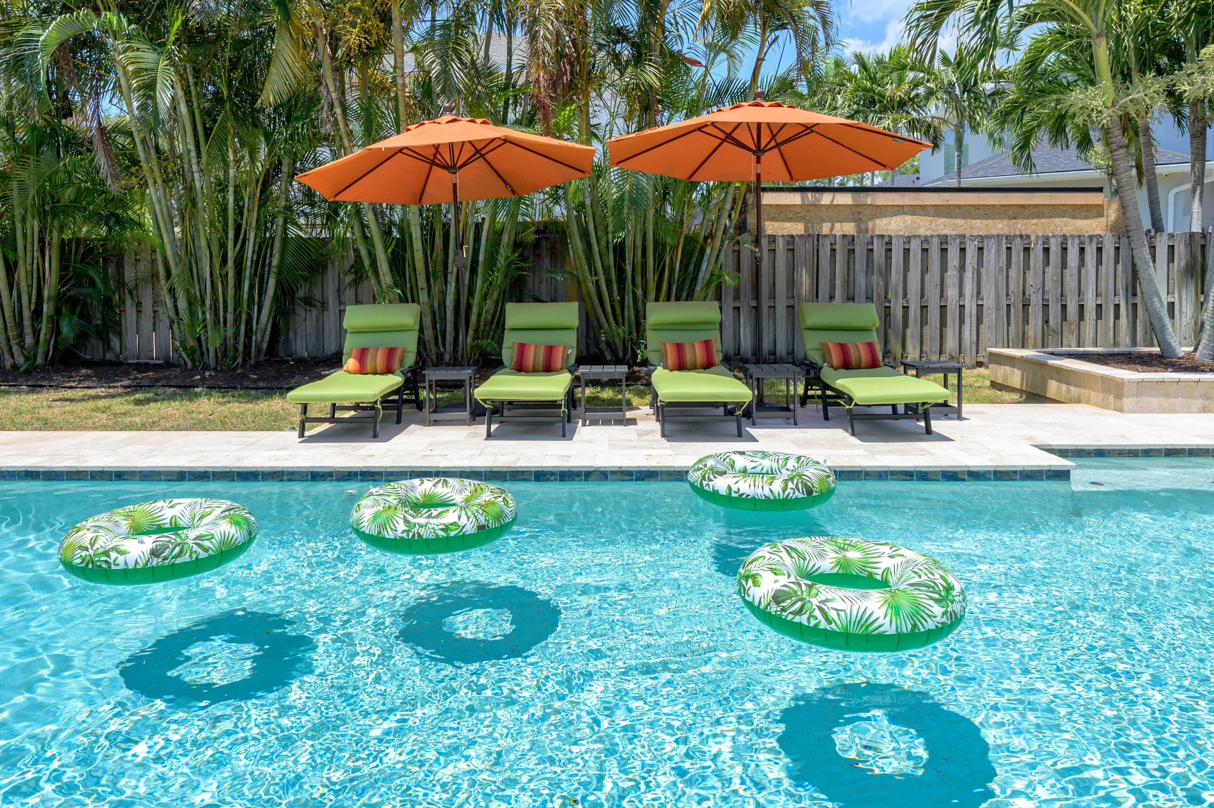 Swimming pool with four floating inflatable rings with tropical leaf patterns, four green lounge chairs with red and yellow striped pillows, and two orange umbrellas on a poolside deck with tropical plants and a wooden fence in the background.