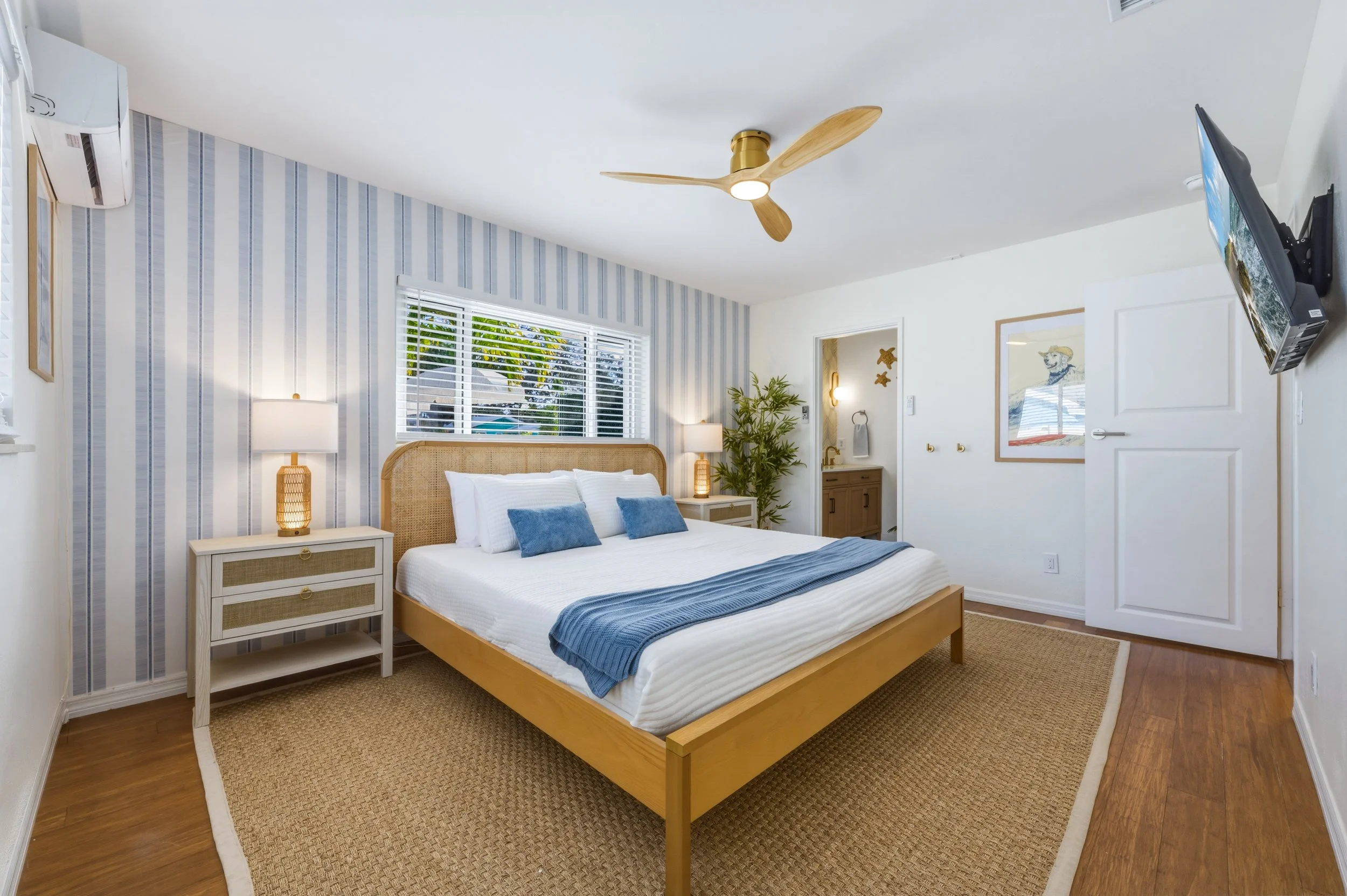 A cozy bedroom with a made bed, blue and white bedding, rattan headboard, and matching nightstands with lamps. There is a window with white blinds, a wall-mounted TV, a ceiling fan, and a small bathroom area with a plant and artwork.