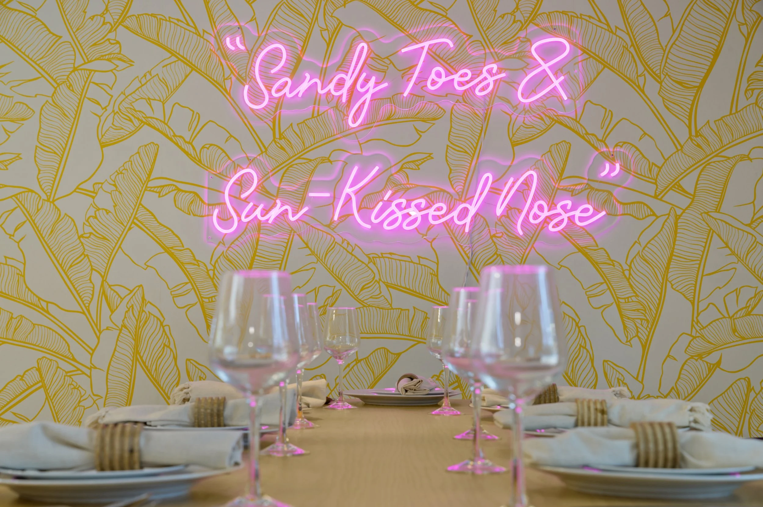 A dining table set for a meal with white napkins secured with gold napkin rings, placed on white plates, and surrounded by tall wine glasses with pink stems. In the background, a wall decorated with gold outlines of banana leaves features a neon pink