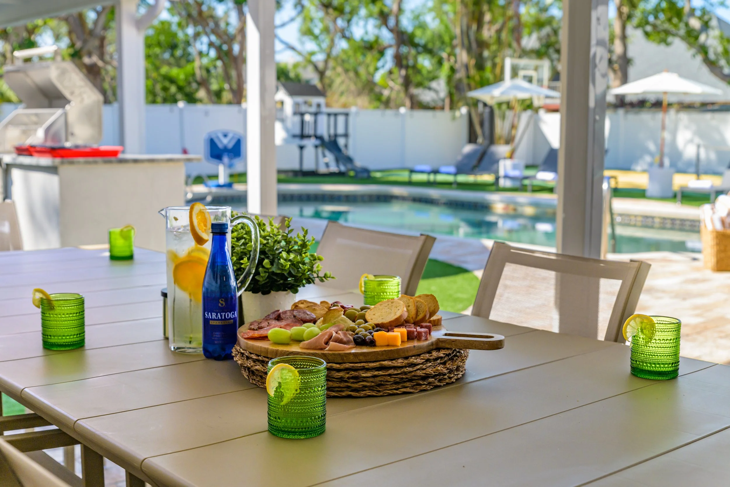 Outdoor patio with a dining table set for a meal, featuring a wooden cheese platter, a pitcher of lemon water, green glasses with lemon slices, a potted plant, and a pool in the background.