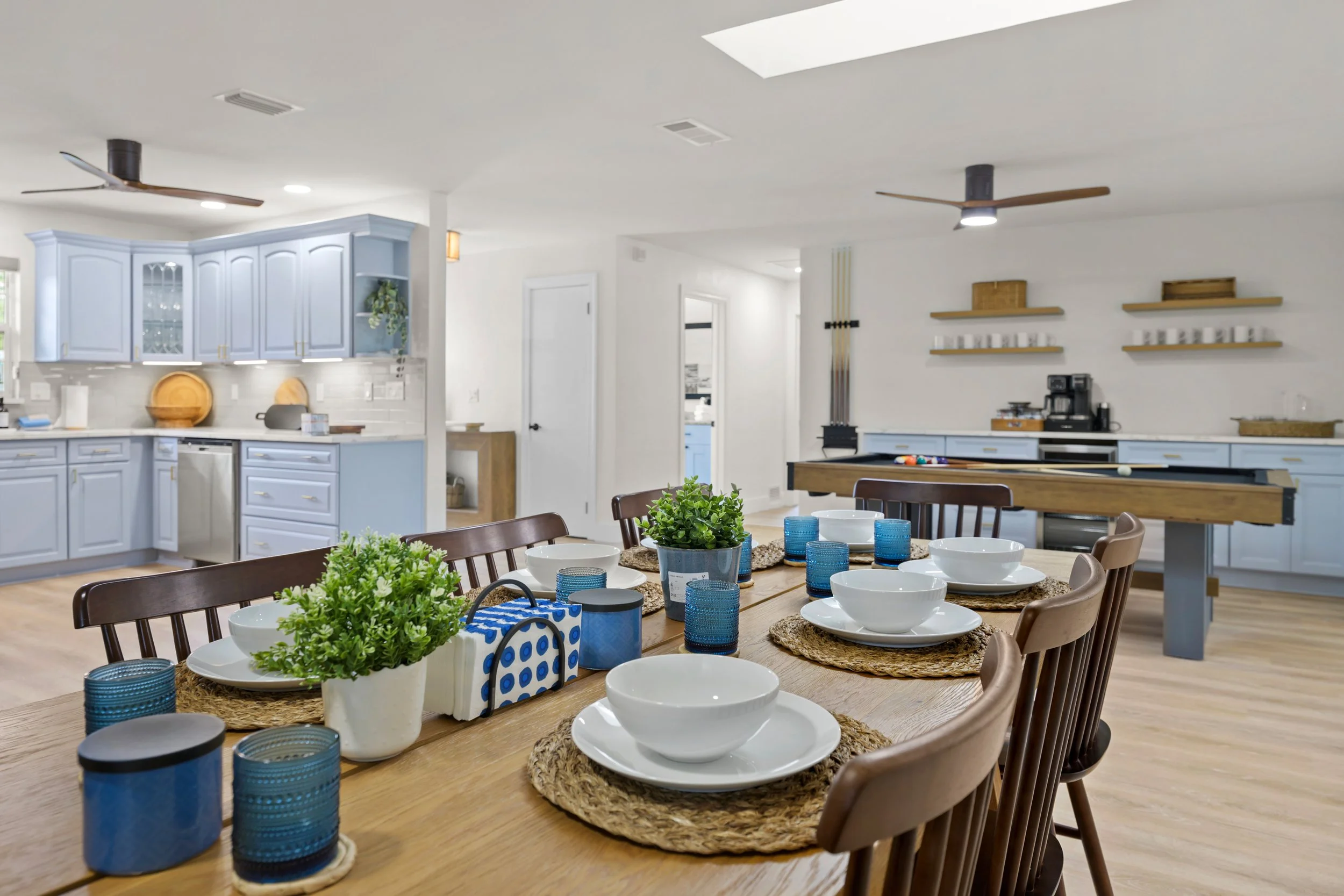 Dining room with a wooden table set with white bowls, plates, blue glasses, woven placemats, and potted greenery, with a kitchen in the background featuring blue cabinets and a pool table.
