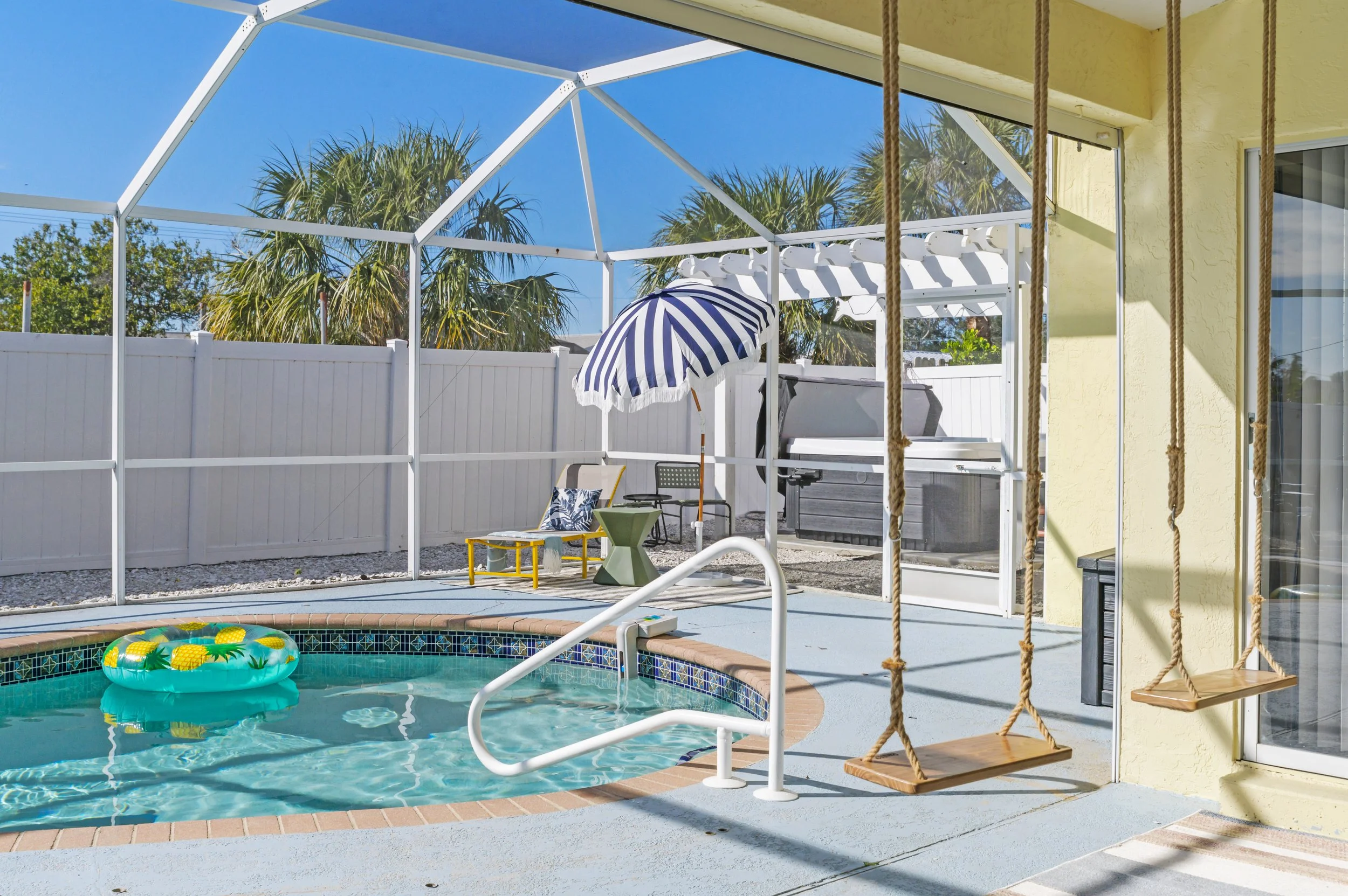 Enclosed backyard pool area with a small swimming pool featuring a pineapple float, a striped patio umbrella, lounge chair with pillow, barbecue grill, and hanging wooden swings. Palm trees and a clear blue sky are visible beyond the white fenced enc