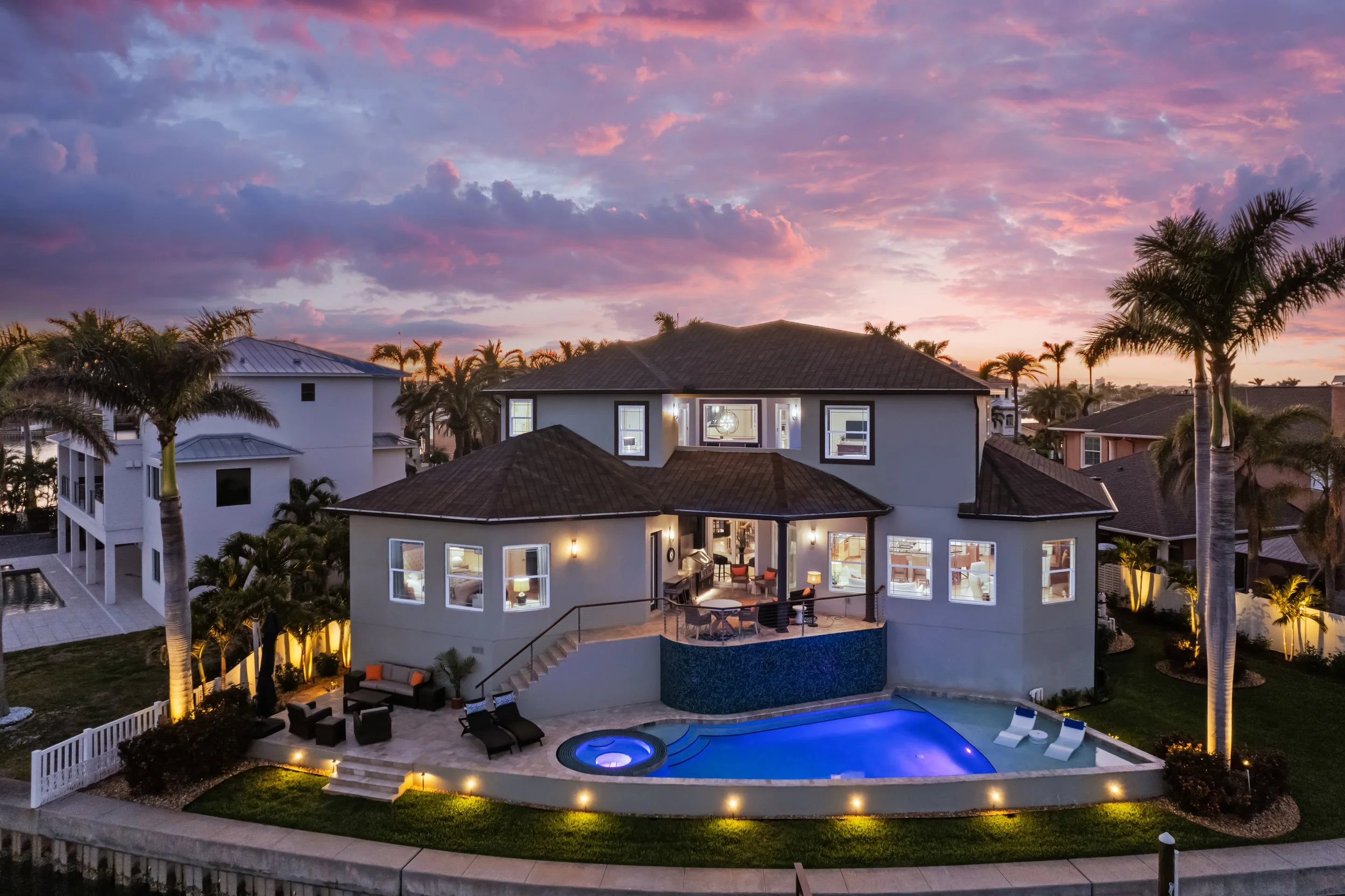Luxury house with swimming pool, outdoor furniture, surrounded by palm trees at sunset.