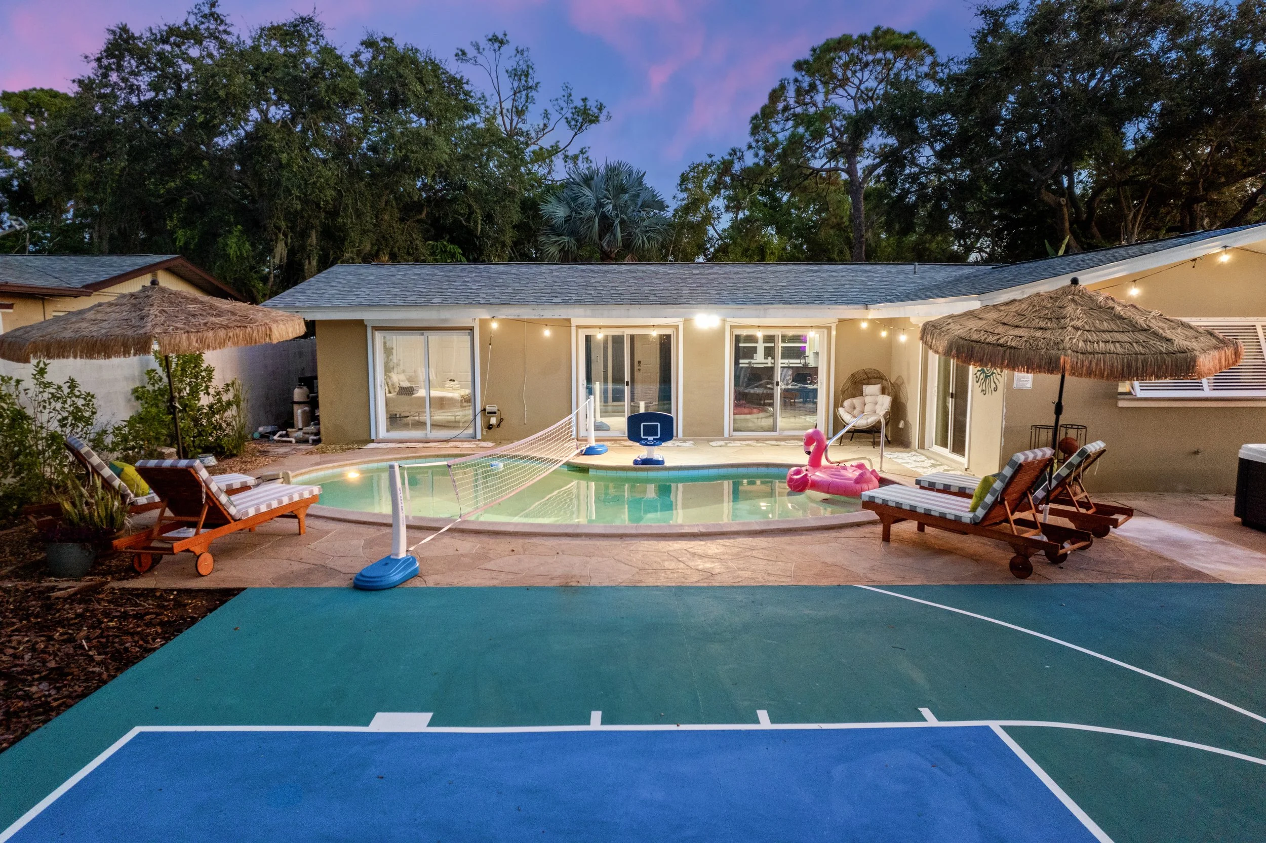 Backyard with swimming pool, lounge chairs, umbrellas, and a basketball hoop, during dusk with outdoor lighting and trees in the background.