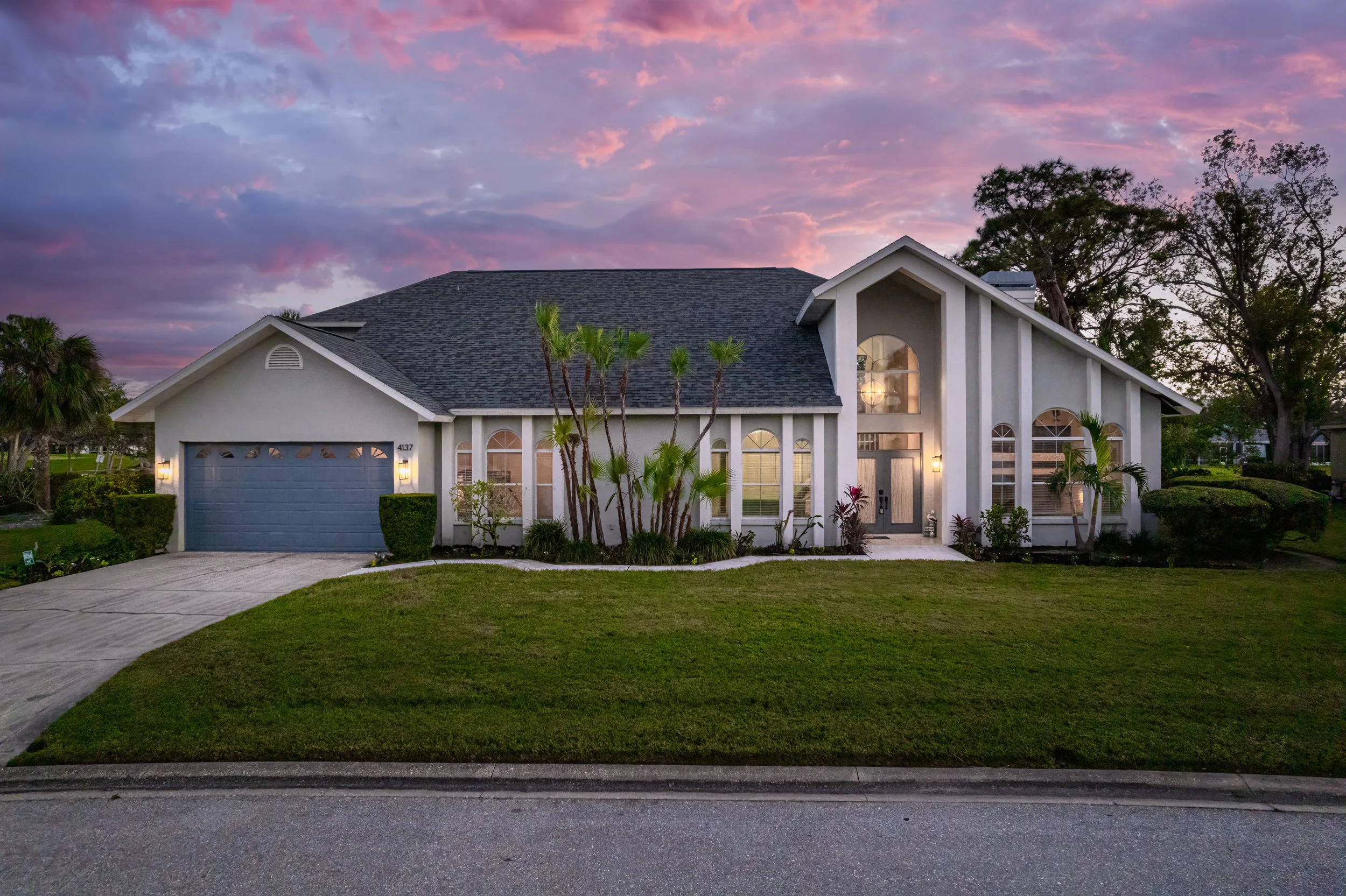 A modern suburban house with a grey roof and white exterior walls, illuminated by exterior lights at dusk, surrounded by a well-maintained lawn and garden with palm trees.