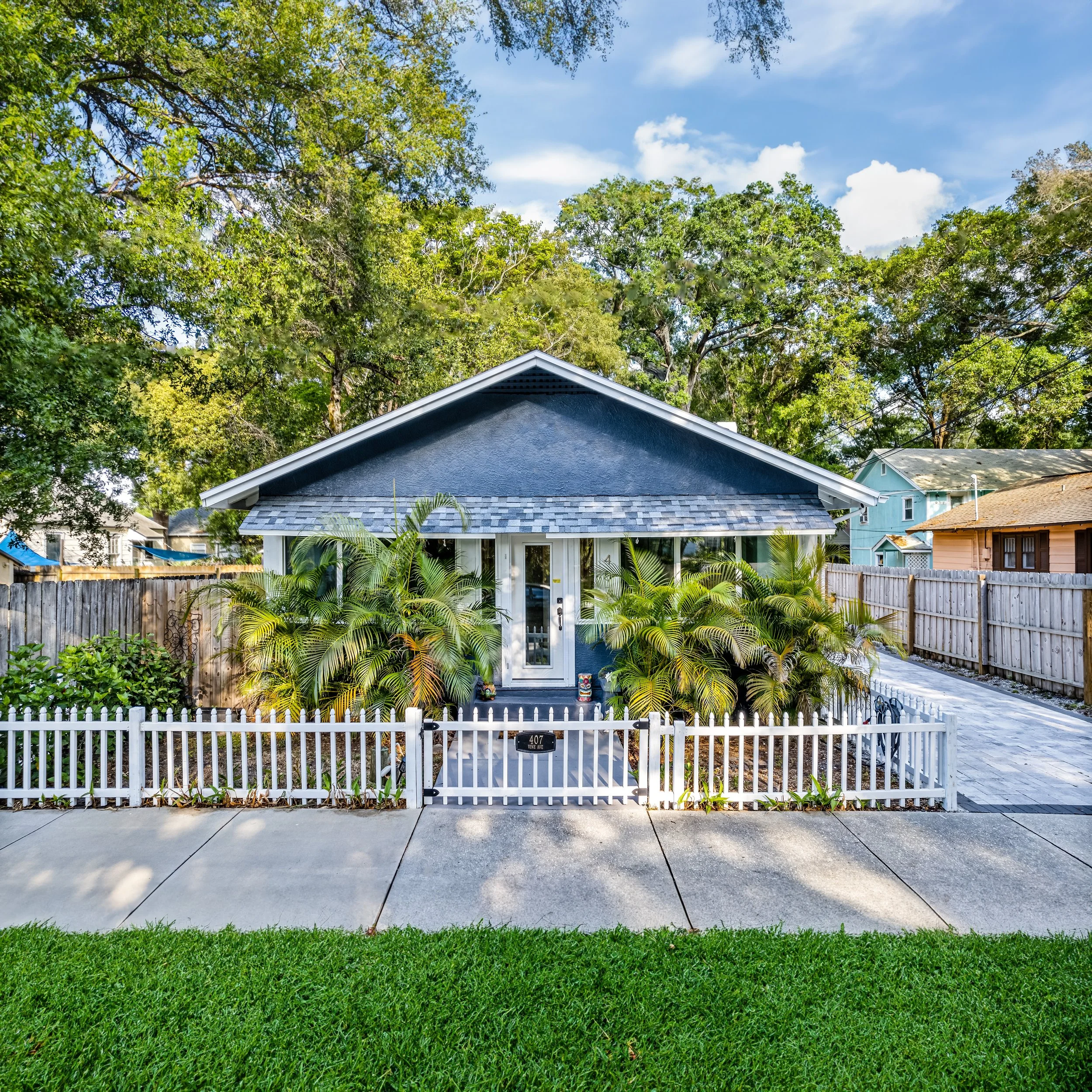 Small blue bungalow house with a white picket fence, surrounded by trees and landscaping, under a clear blue sky.