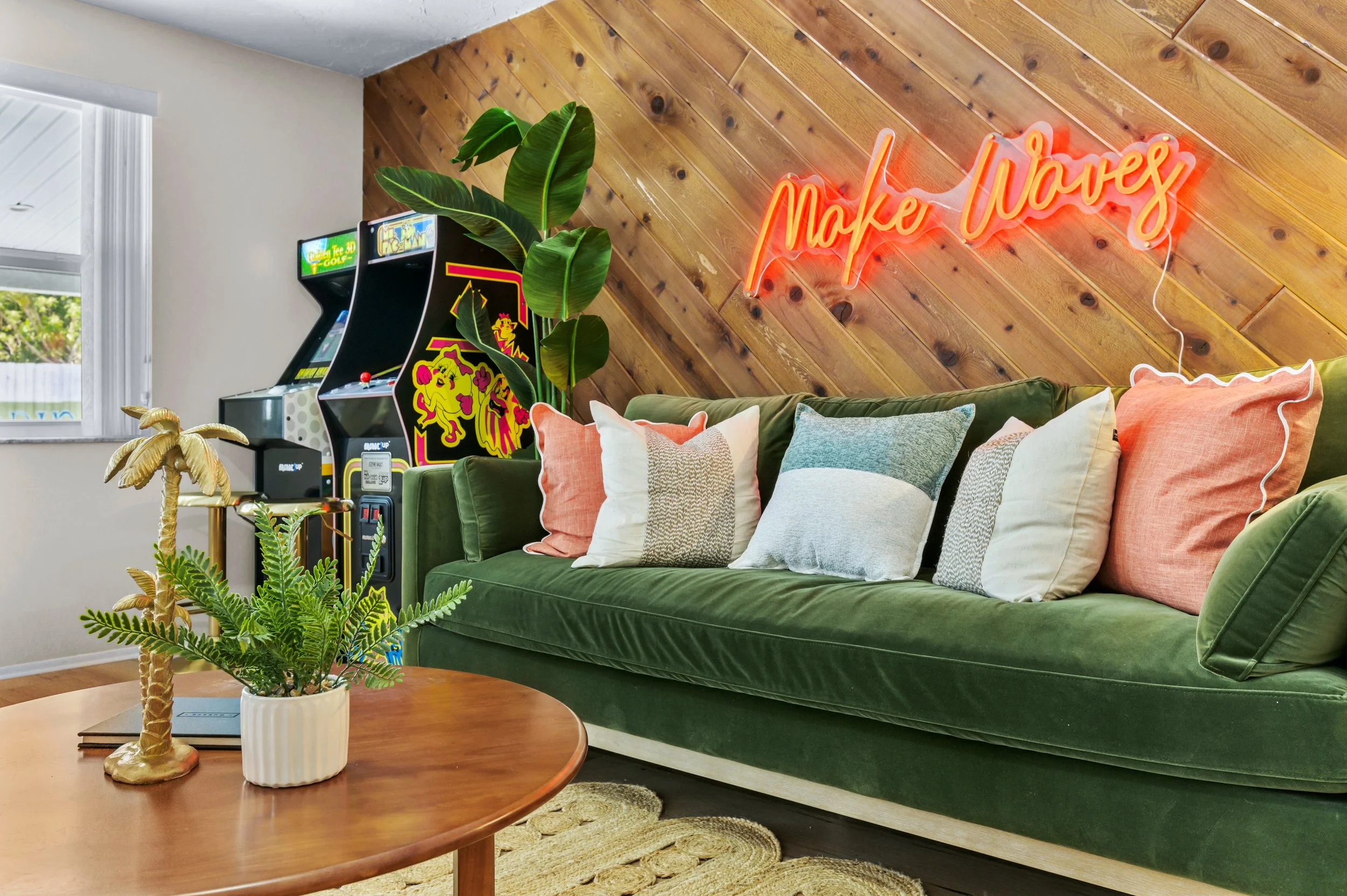 A cozy living room with a green velvet couch adorned with pillows in pastel colors, a wooden coffee table with a decorative palm tree and fern in a white pot, a wooden paneled wall with a neon sign that says 'Make Waves,' an arcade game, and a large