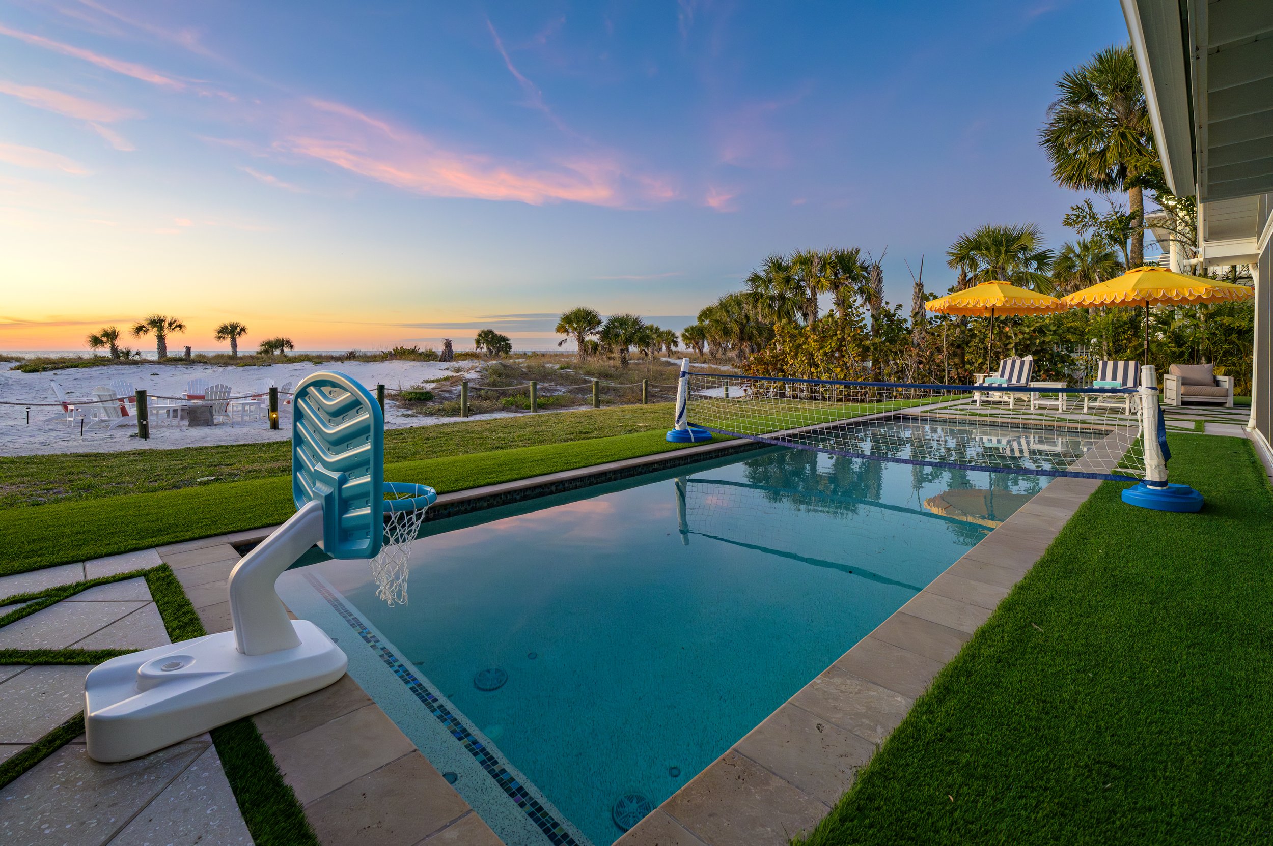 A backyard swimming pool with a volleyball net and a basketball hoop, surrounded by a grassy area. In the background, a beach with white sand and palm trees is visible, along with a sunset sky. There are also outdoor seating and yellow umbrellas on t