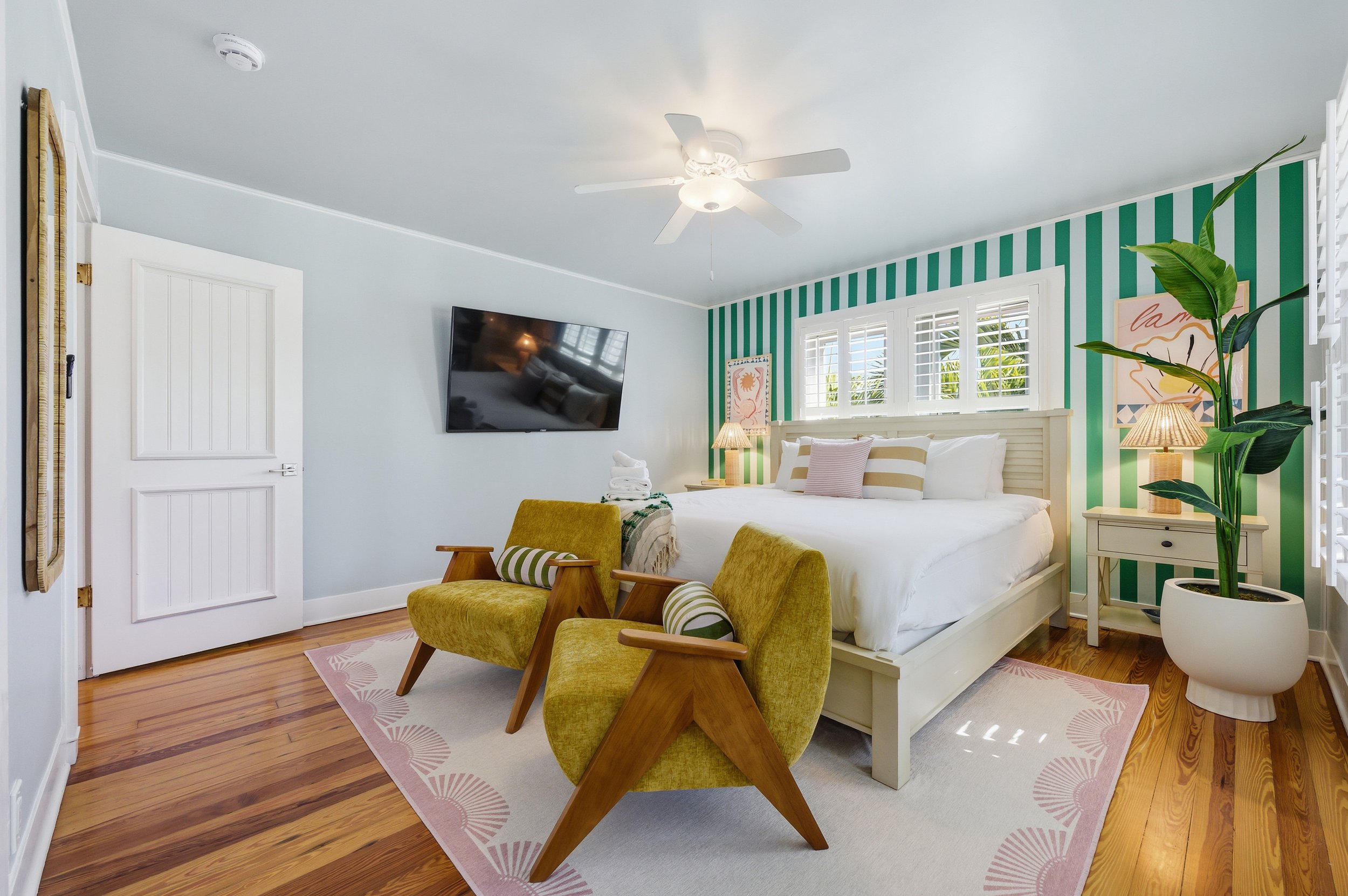 Bright bedroom with light blue and green striped wall behind a white bed, two yellow armchairs with wooden arms, a potted plant, and a TV on the wall.