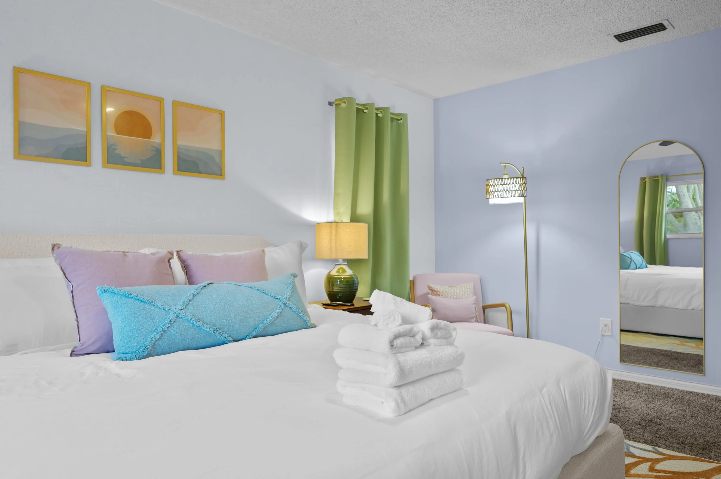 Cozy bedroom with a neatly made bed, pastel pillows, a green lamp, a pink chair, a standing lamp, green curtains, and wall art of a sunset over water. A large mirror reflects part of the room.