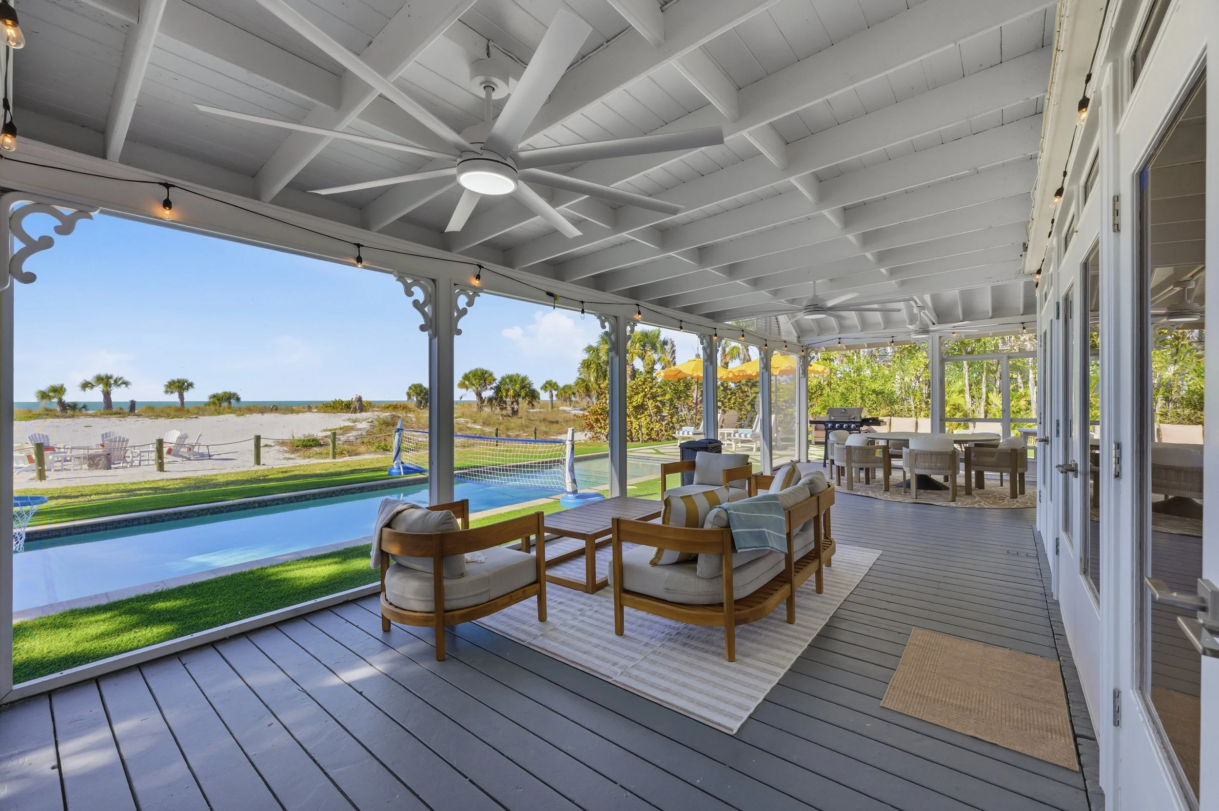 Screened-in porch with wooden flooring, ceiling fan, and comfortable seating area overlooking a backyard with a pool, volleyball net, and sandy beach with palm trees in the distance.