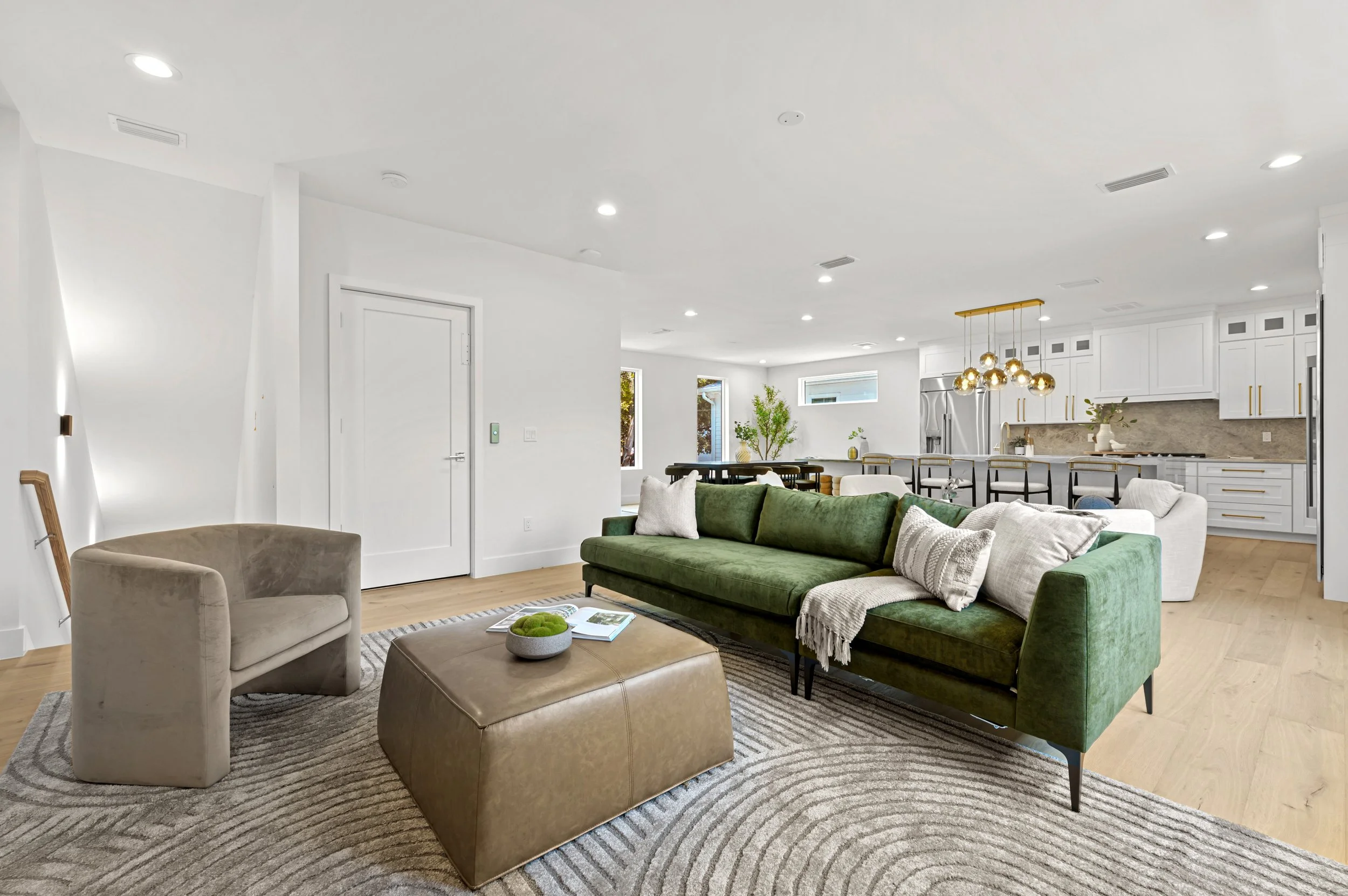Open-concept living room and kitchen with white walls, hardwood floors, green and beige sofas, area rug, and modern kitchen cabinetry and appliances.