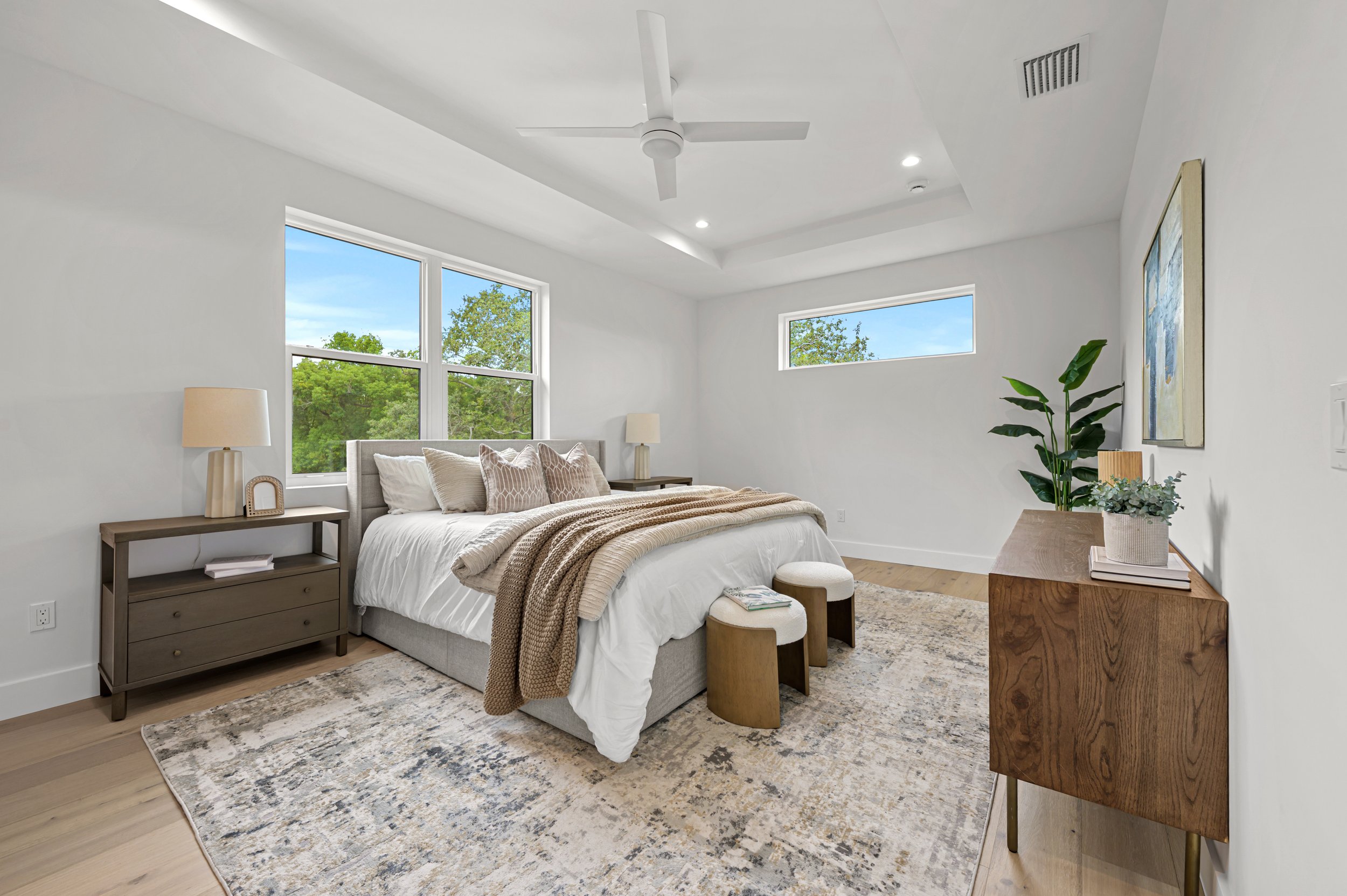 A modern bedroom with a white bed, beige throw blanket, and multiple pillows. There are two wooden nightstands with lamps, a wooden dresser with a plant and books, and a large window showing greenery outside. The room has light-colored hardwood floor