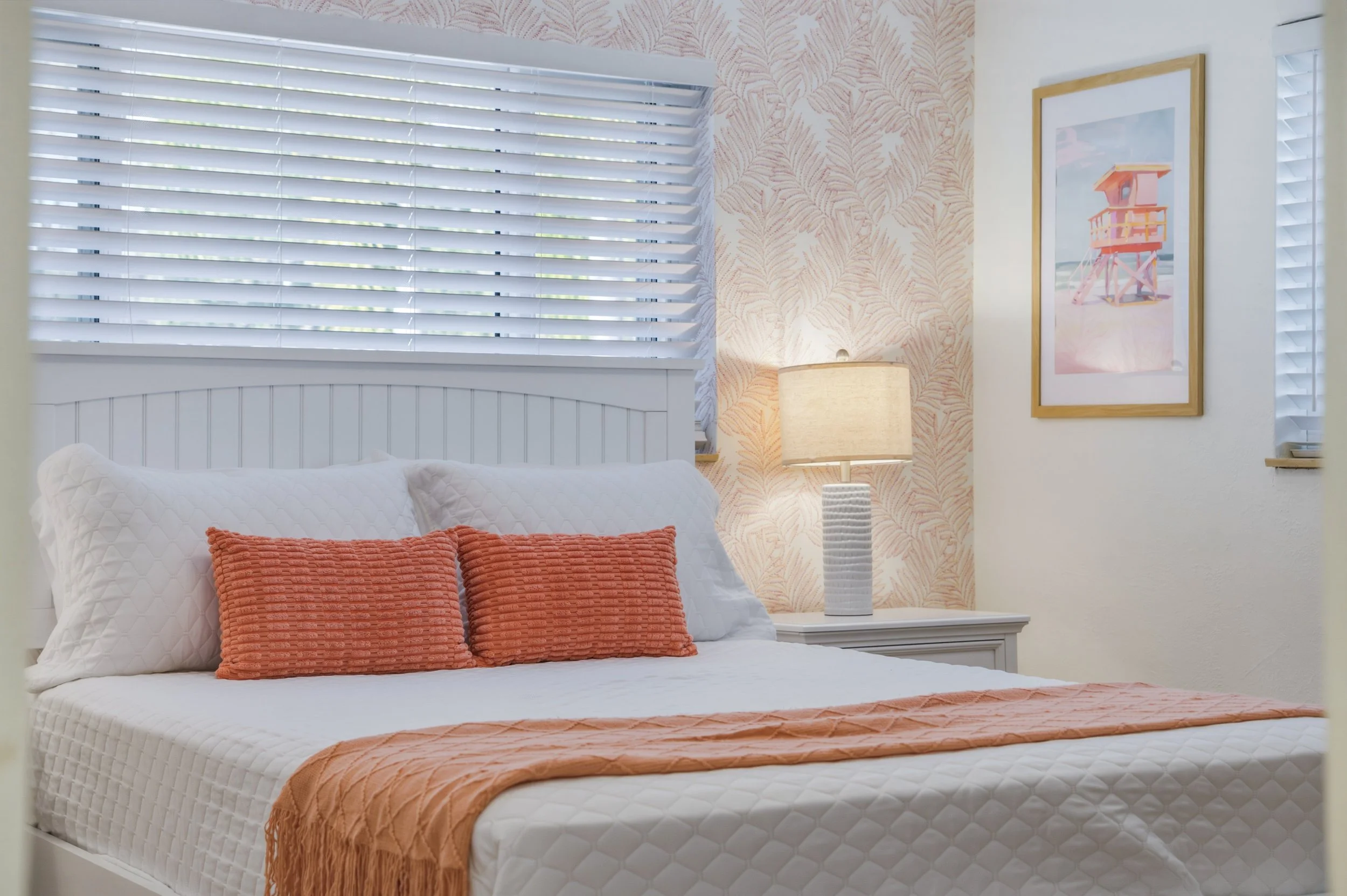 A neatly made bedroom with white bedding, two orange throw pillows, and a matching orange blanket. There is a white nightstand with a textured white lamp. The room has light-colored wallpaper with a leaf pattern and framed coastal artwork on the wall