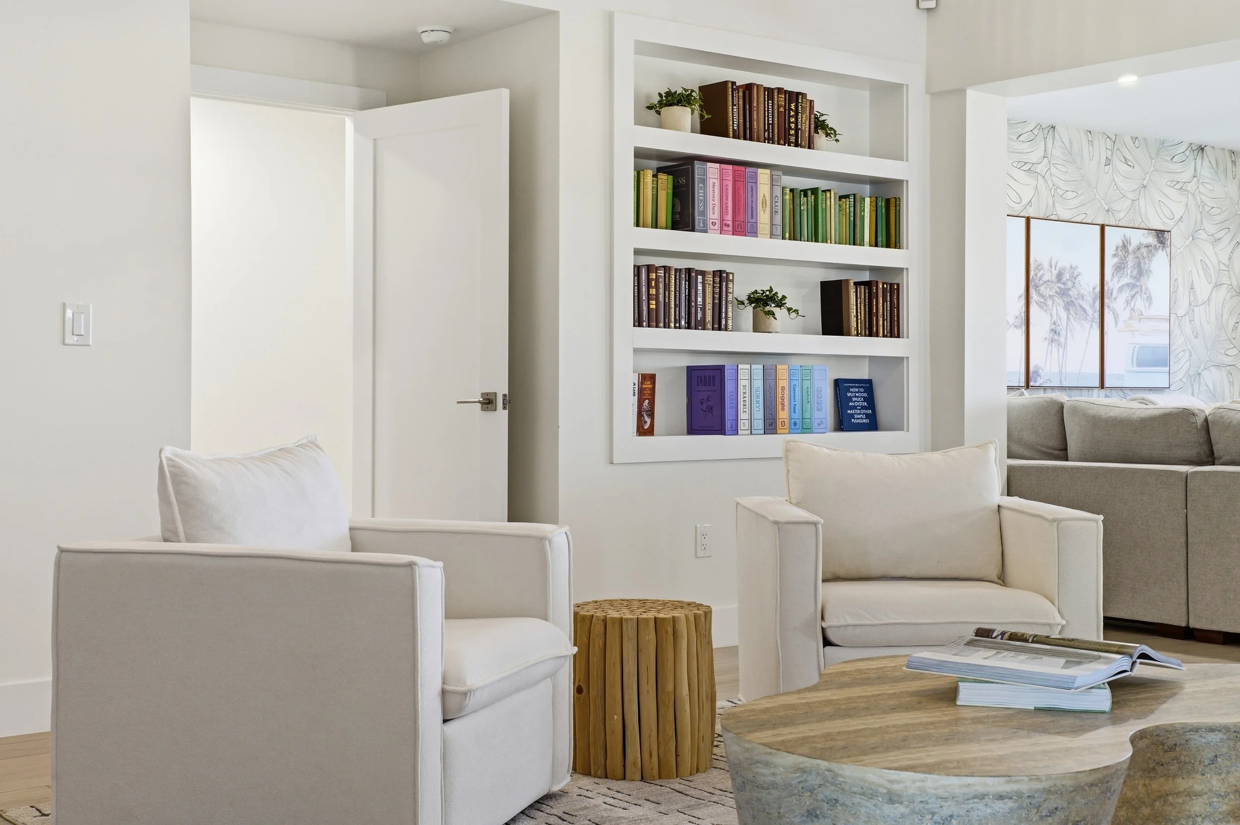 Living room with two white armchairs, a wooden side table, a beige couch, a white built-in bookshelf filled with books and plants, and a stone coffee table with open magazines, near a window with palm tree views.
