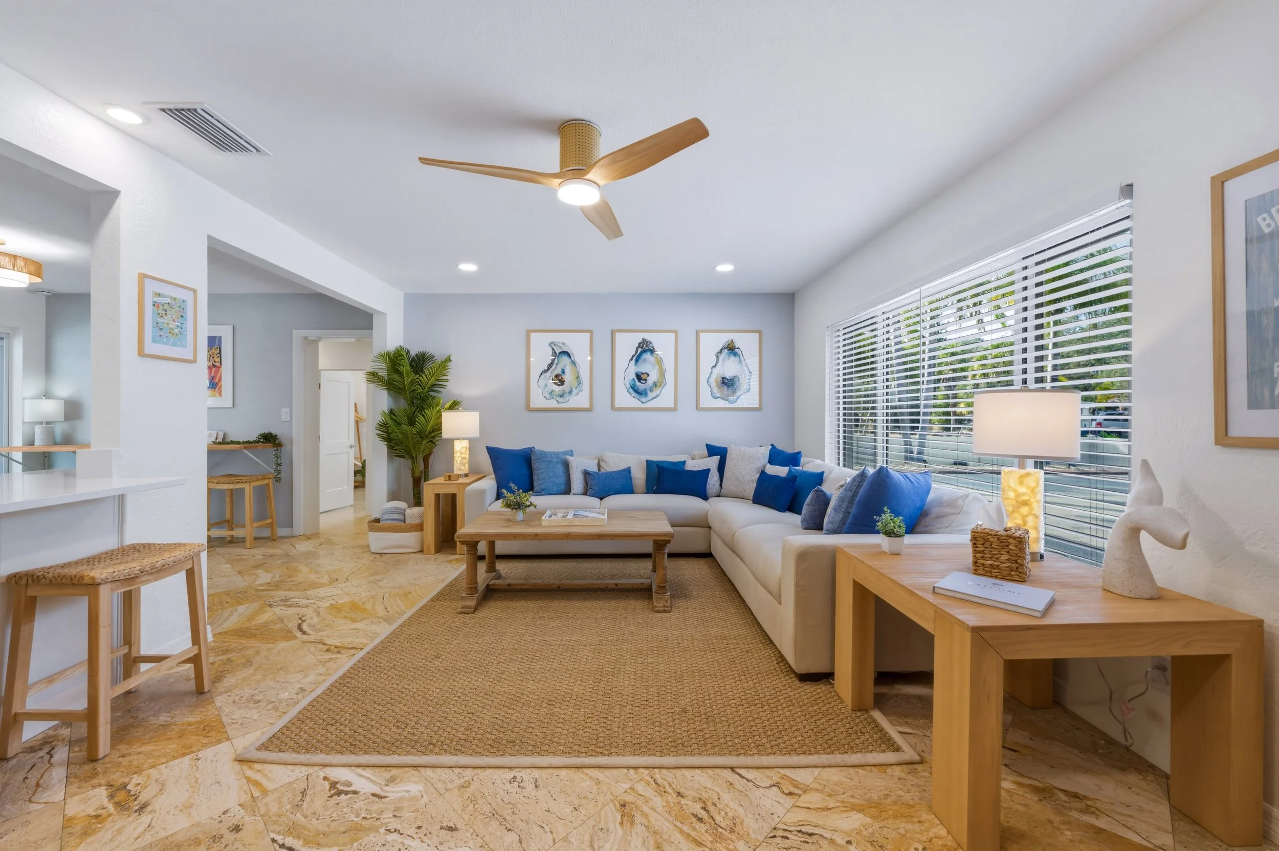 Bright living room with white sectional sofa adorned with various blue and gray pillows, a wooden coffee table, side tables, a large window with white blinds, decorative artwork, and indoor plants.