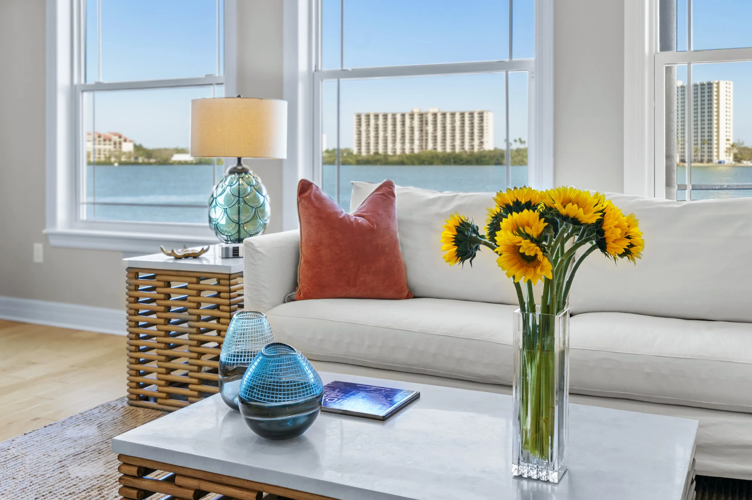 Bright living room with white sofa, yellow sunflower bouquet, orange pillow, rattan side table with table lamp, decorative blue vases, and large windows with water and high-rise buildings outside.