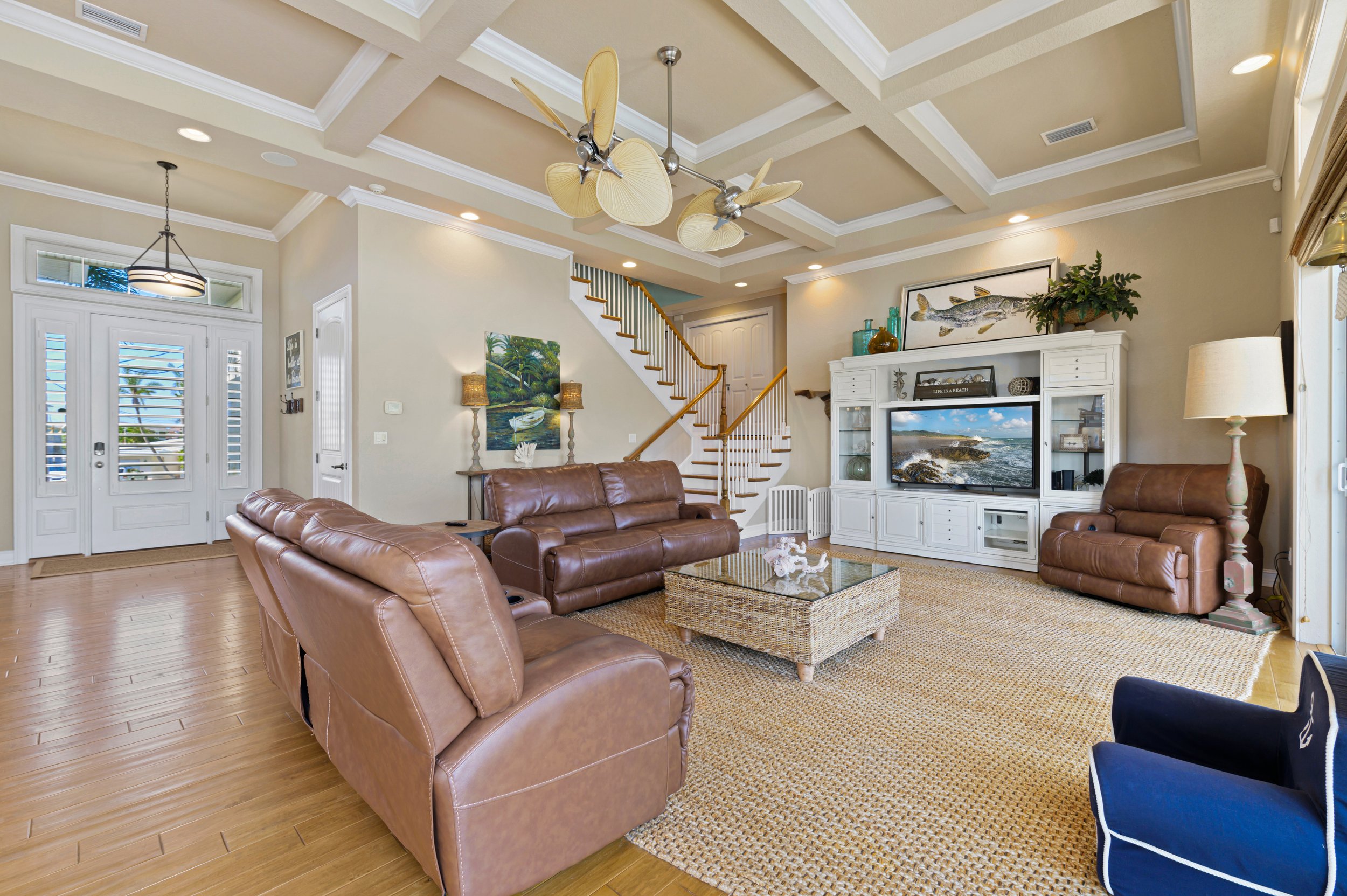 Spacious living room with brown leather sofas, wicker coffee table, large television on a white media cabinet, and a ceiling fan. The room features a staircase, hardwood flooring, and beige walls, with decorative elements like paintings and lamps.