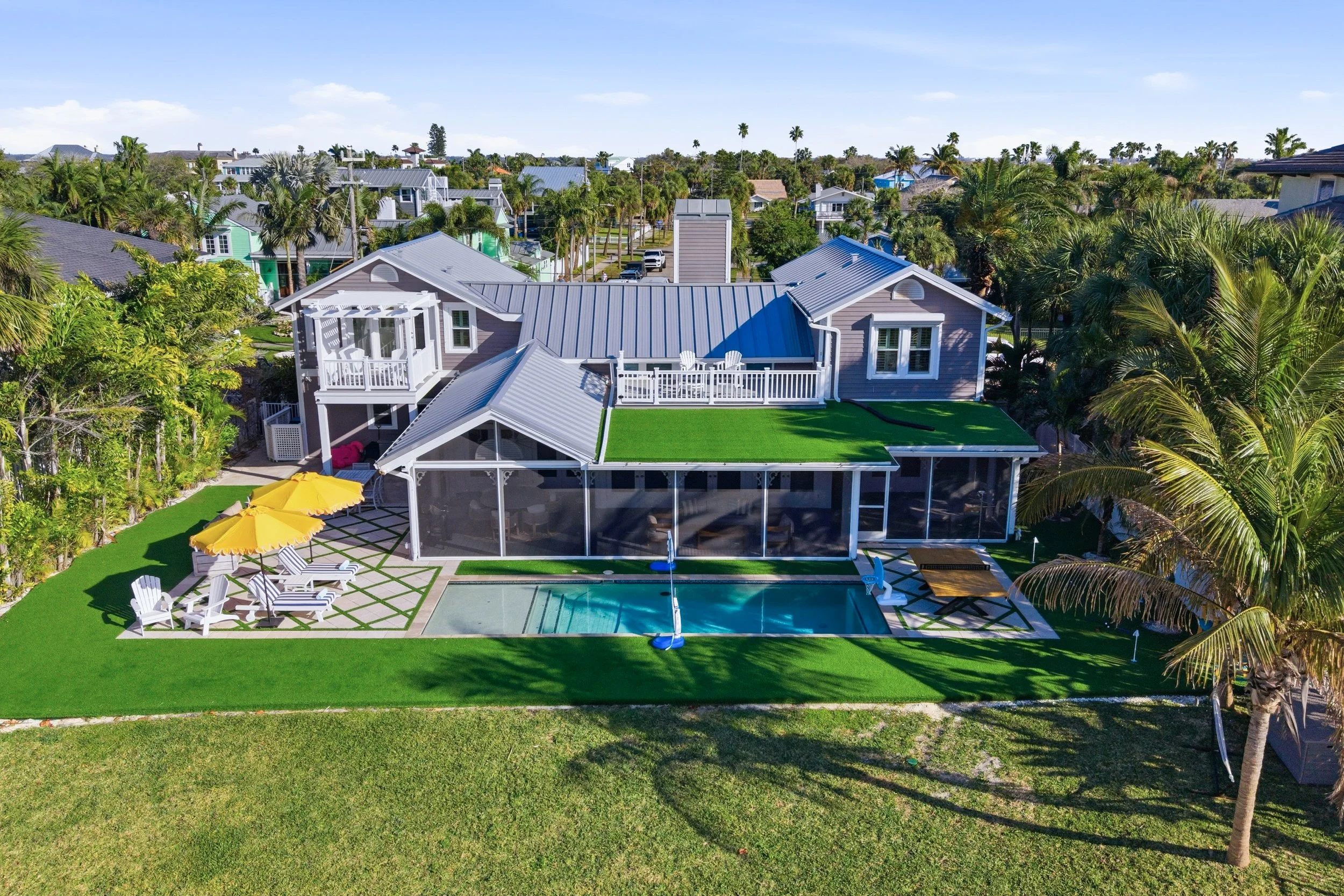 A large backyard with a swimming pool, outdoor furniture, and a screened porch connected to a light blue house with a second-floor balcony. The yard is landscaped with green grass, palm trees, and a yellow patio umbrella.