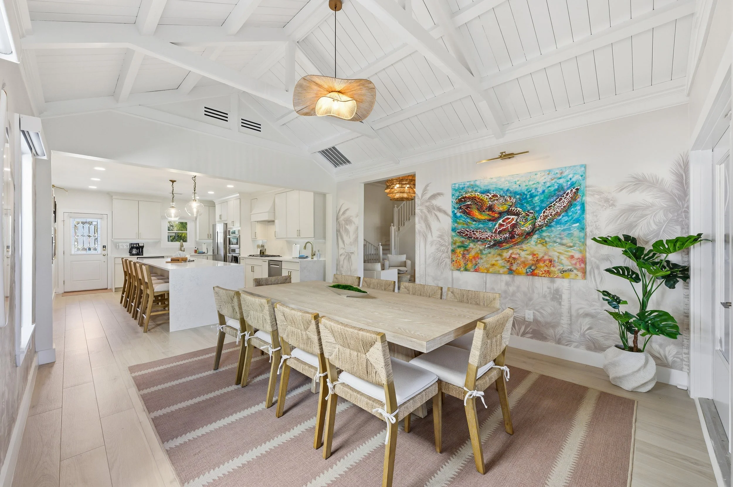 Open-concept dining area with a large wooden table and ten chairs, beige rug, tropical-themed wall art, green potted plant, and view into a bright white kitchen with bar stools and modern appliances.