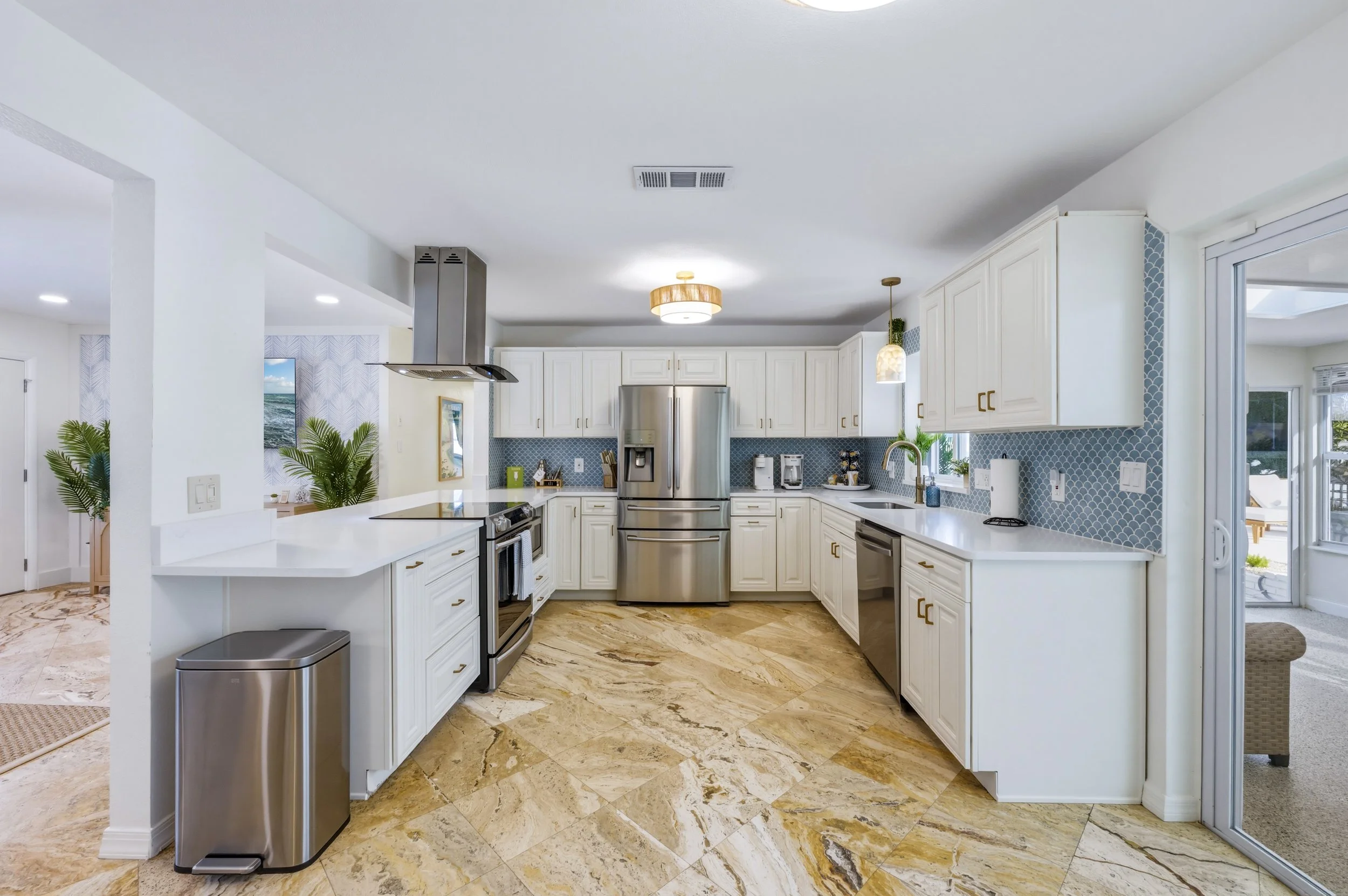 Modern kitchen with white cabinets, stainless steel appliances, blue tile backsplash, beige marble floor, and a sliding glass door leading outside.