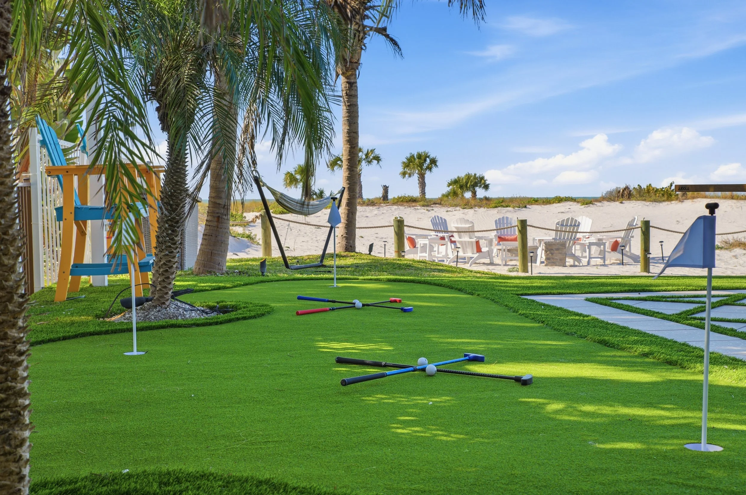 Mini golf course with palm trees, putting greens, golf clubs, and flags, bordered by sand dunes and beach chairs in the background, under a clear blue sky.