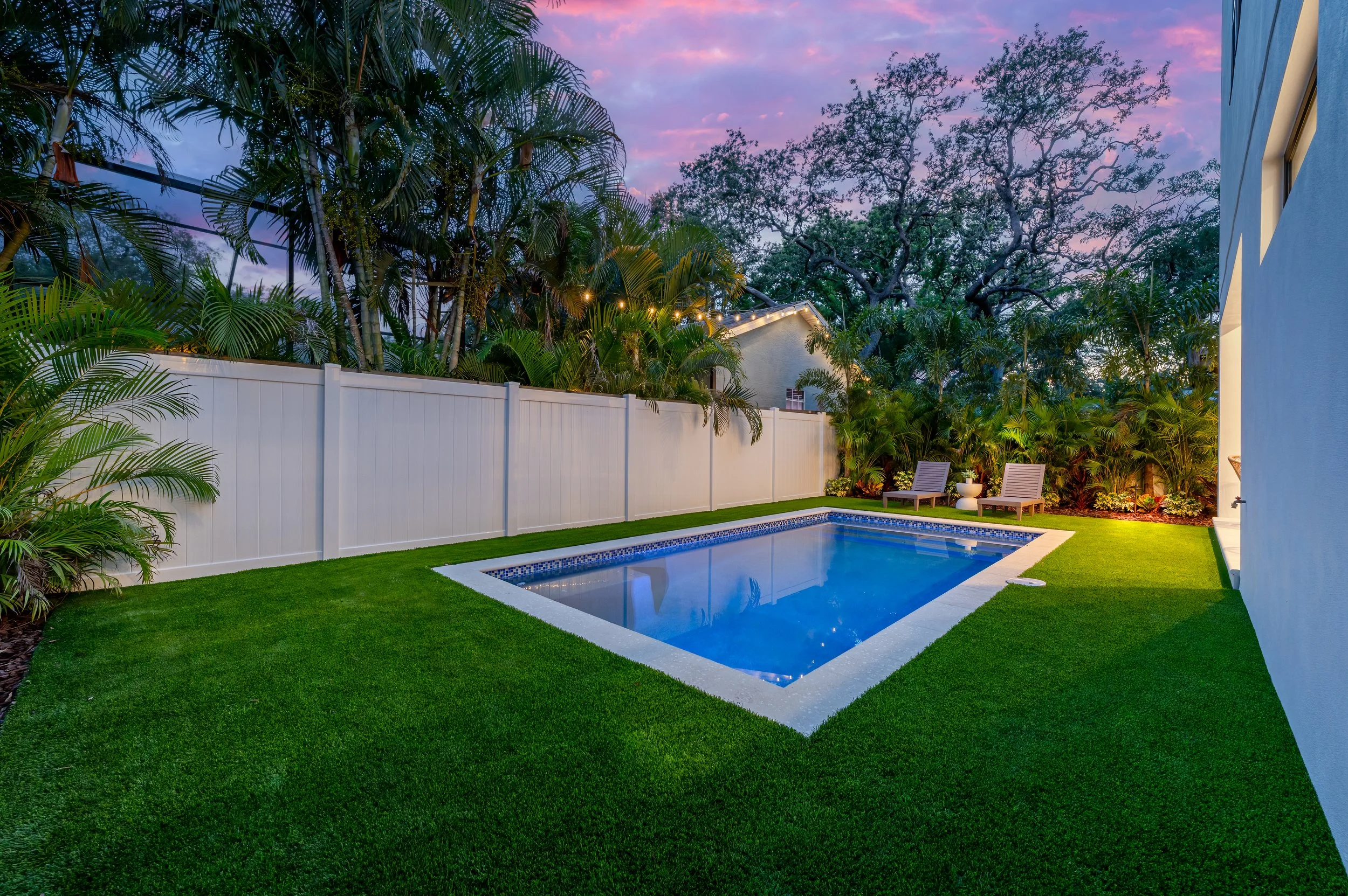 A backyard with a small rectangular pool, surrounded by a well-manicured green lawn and white fencing. There are two lounge chairs and a small table with a plant in the corner, and tall trees and lush bushes in the background. Twilight sky with pink 