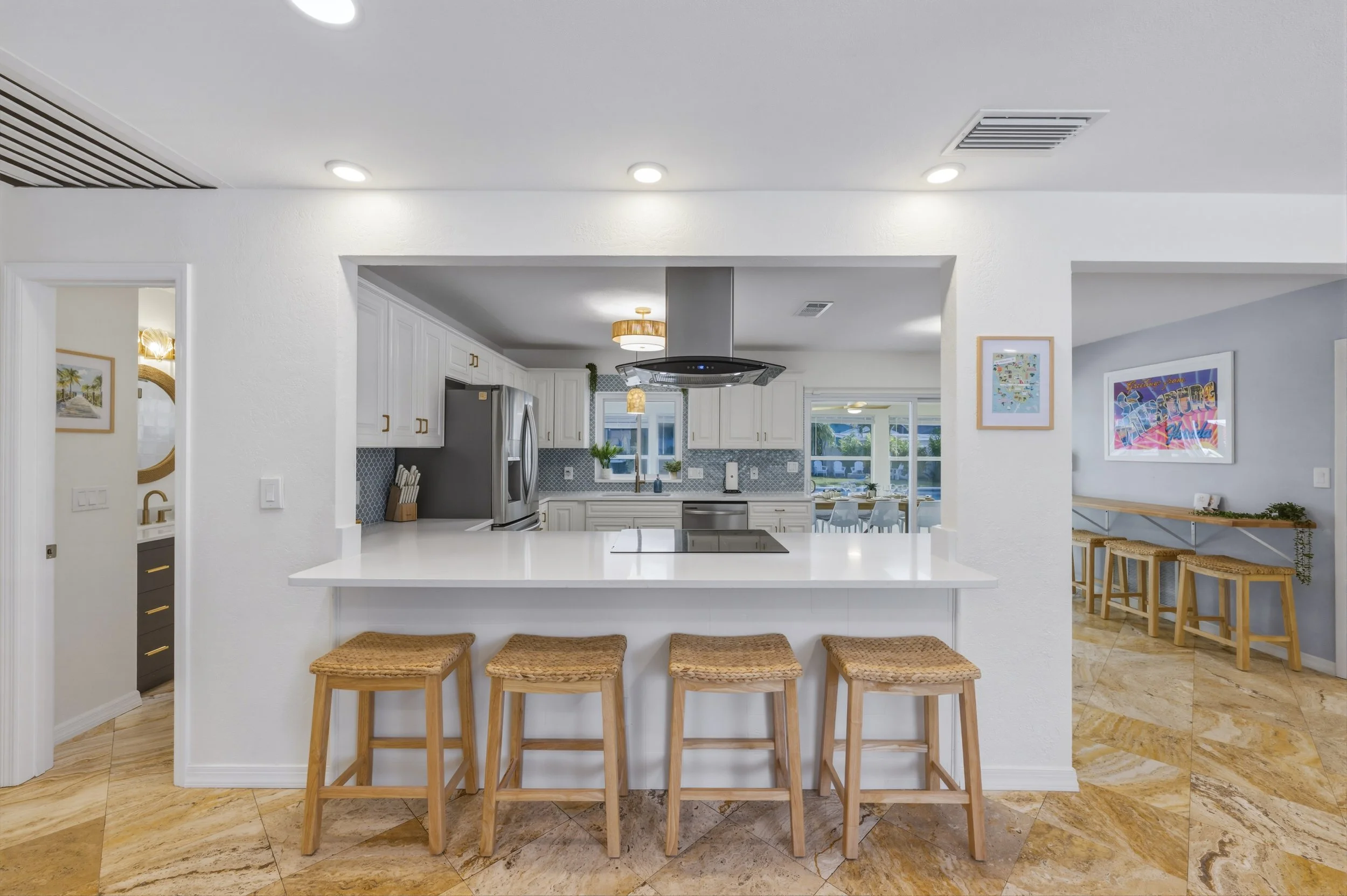 Modern kitchen with white cabinets, stainless steel appliances, and a white island with four rattan stools. There is a view of a dining area and living room through large windows, and artwork decorates the walls. The floor is tan marble tiles.