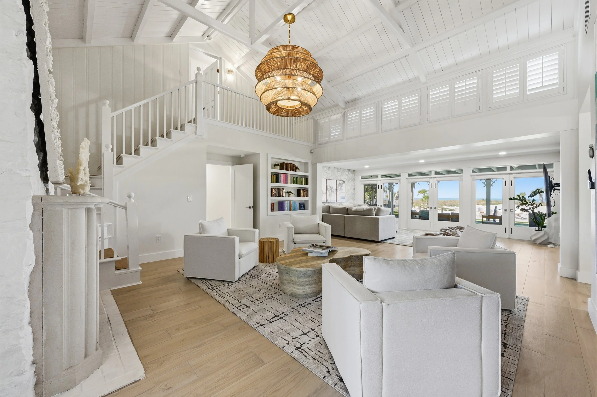 Bright, spacious living room with white walls and ceiling, wooden flooring, and large windows viewing outdoor scenery. Furniture includes white armchairs, a sofa, a stone coffee table, and a built-in bookshelf. A wicker chandelier hangs from the vaul