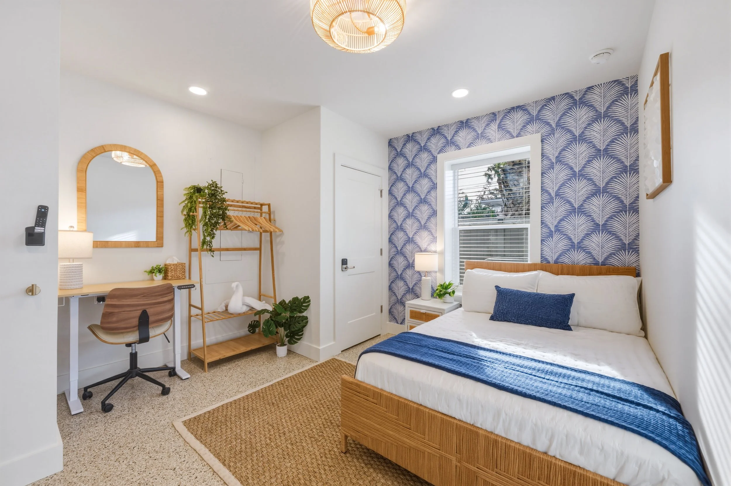 A bright bedroom with a white bed featuring blue pillows and a blue blanket. A window with blinds and a nightstand with a lamp and a plant are beside the bed. The wall behind the bed has a blue-patterned wallpaper. On the left, there is a desk with a