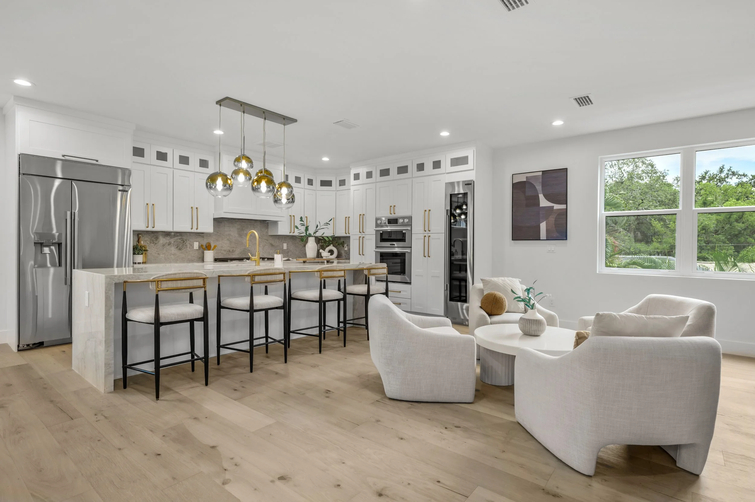 Modern kitchen and living area with white cabinetry, stainless steel refrigerator, island with four barstools, gold fixtures, and a seating area with white chairs and a coffee table, bright natural light through large window.