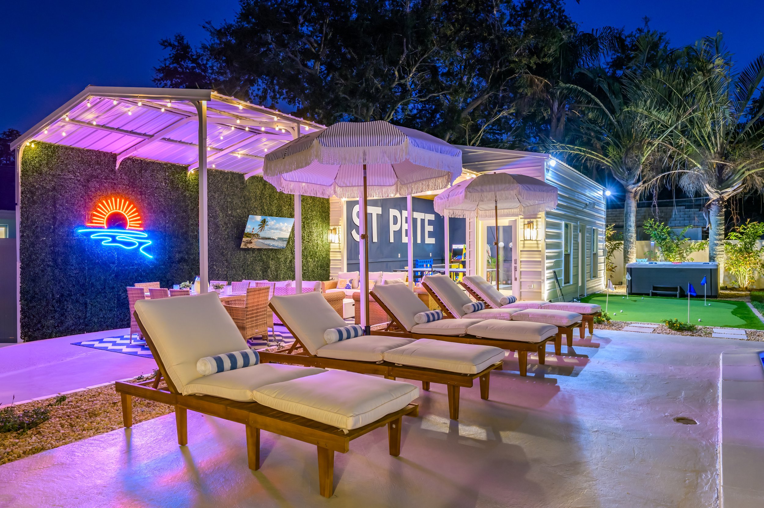 Night view of a backyard with lounge chairs, umbrellas, a small putting green, and decorative neon lights.