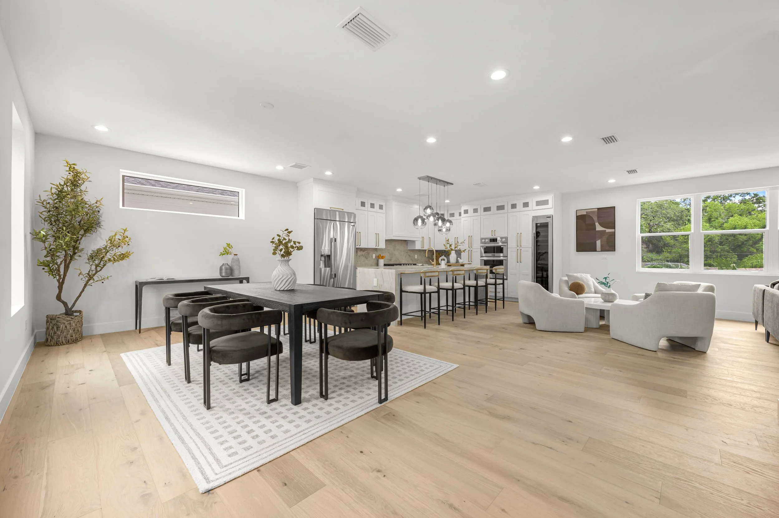 Open-concept living room and kitchen with white walls, light wood flooring, modern furniture, and large windows showing greenery outside.