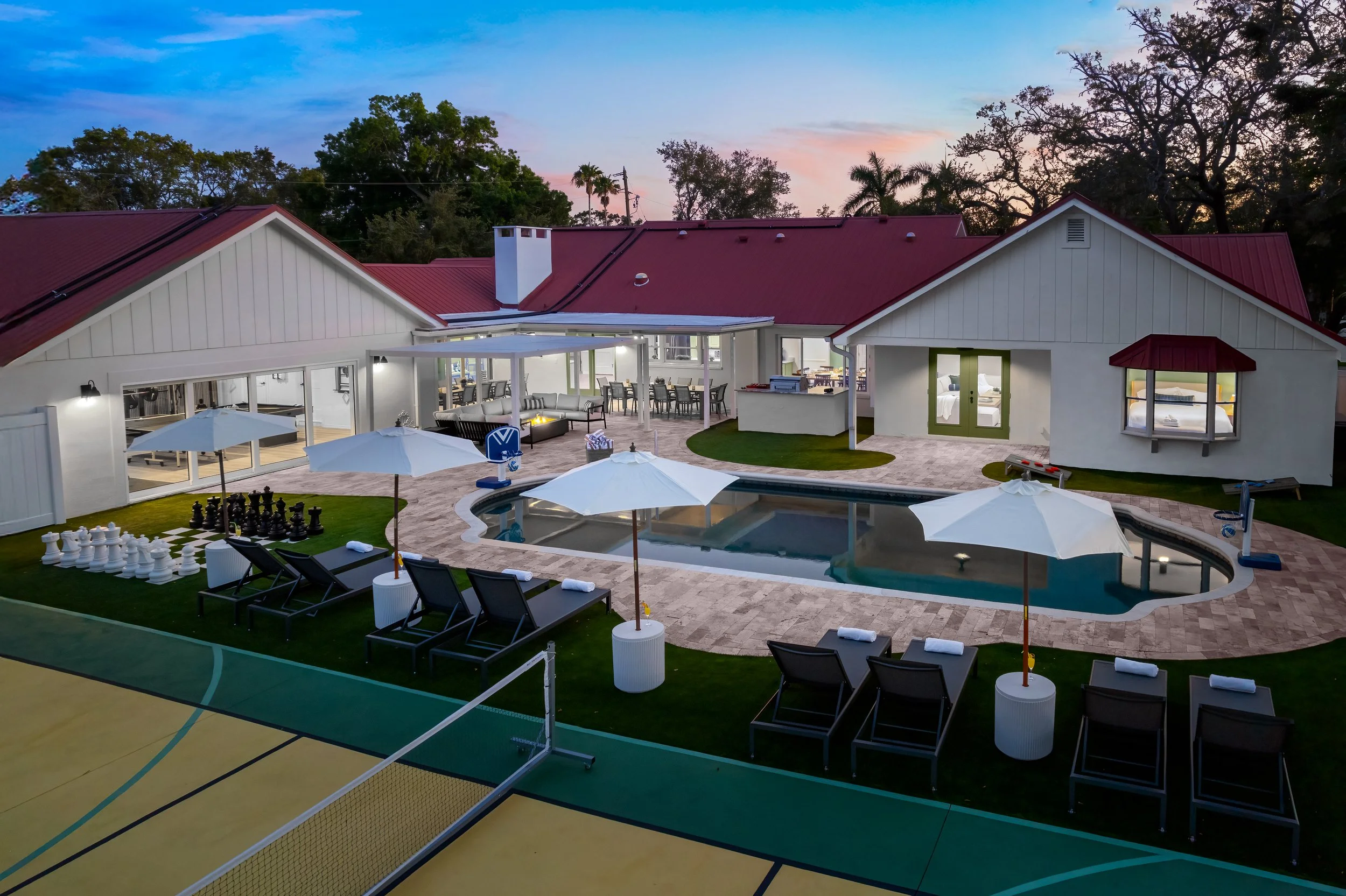 A backyard with a swimming pool, lounge chairs, umbrellas, and a pool basketball hoop, surrounded by a tennis court, with a house in the background featuring a porch and interior lights on during dusk.