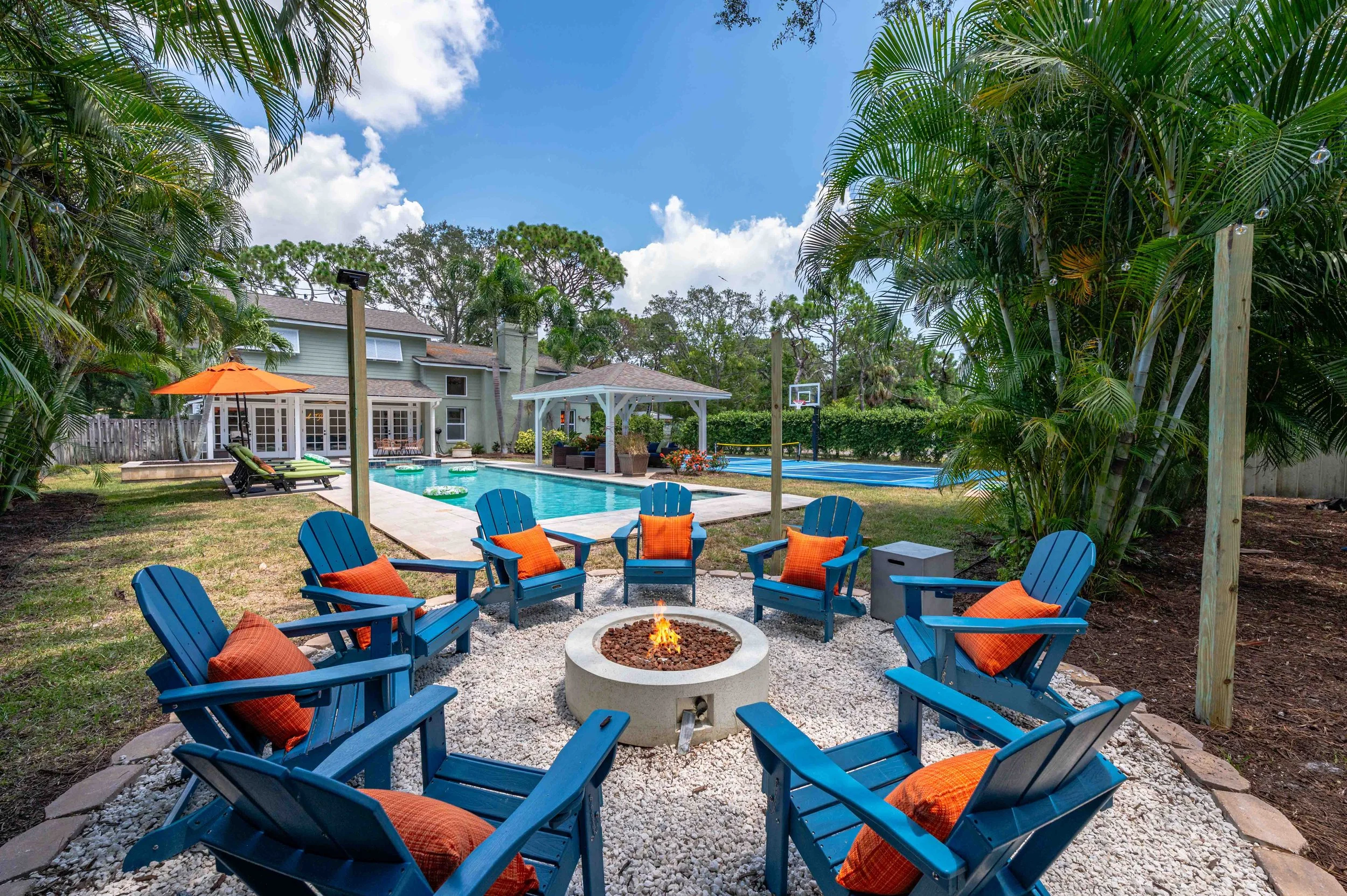Outdoor backyard with a fire pit surrounded by blue Adirondack chairs with orange cushions, a swimming pool, a pavilion, and a separate basketball court surrounded by lush palm trees and greenery under a partly cloudy sky.