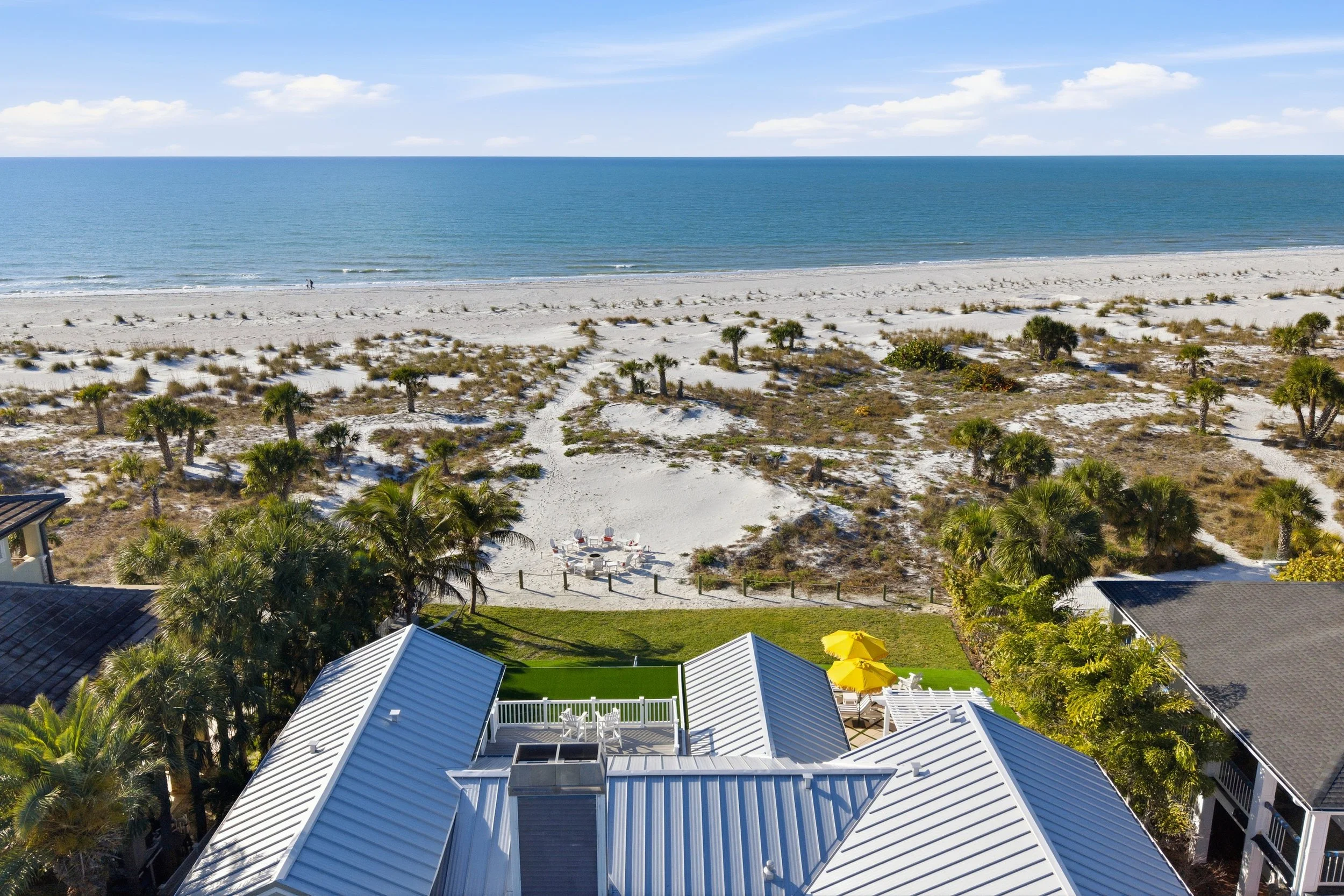 View from above of houses with metal roofs, a backyard with a green lawn, and a patio with yellow umbrellas, overlooking a sandy beach with palm trees and the ocean in the distance under a blue sky.