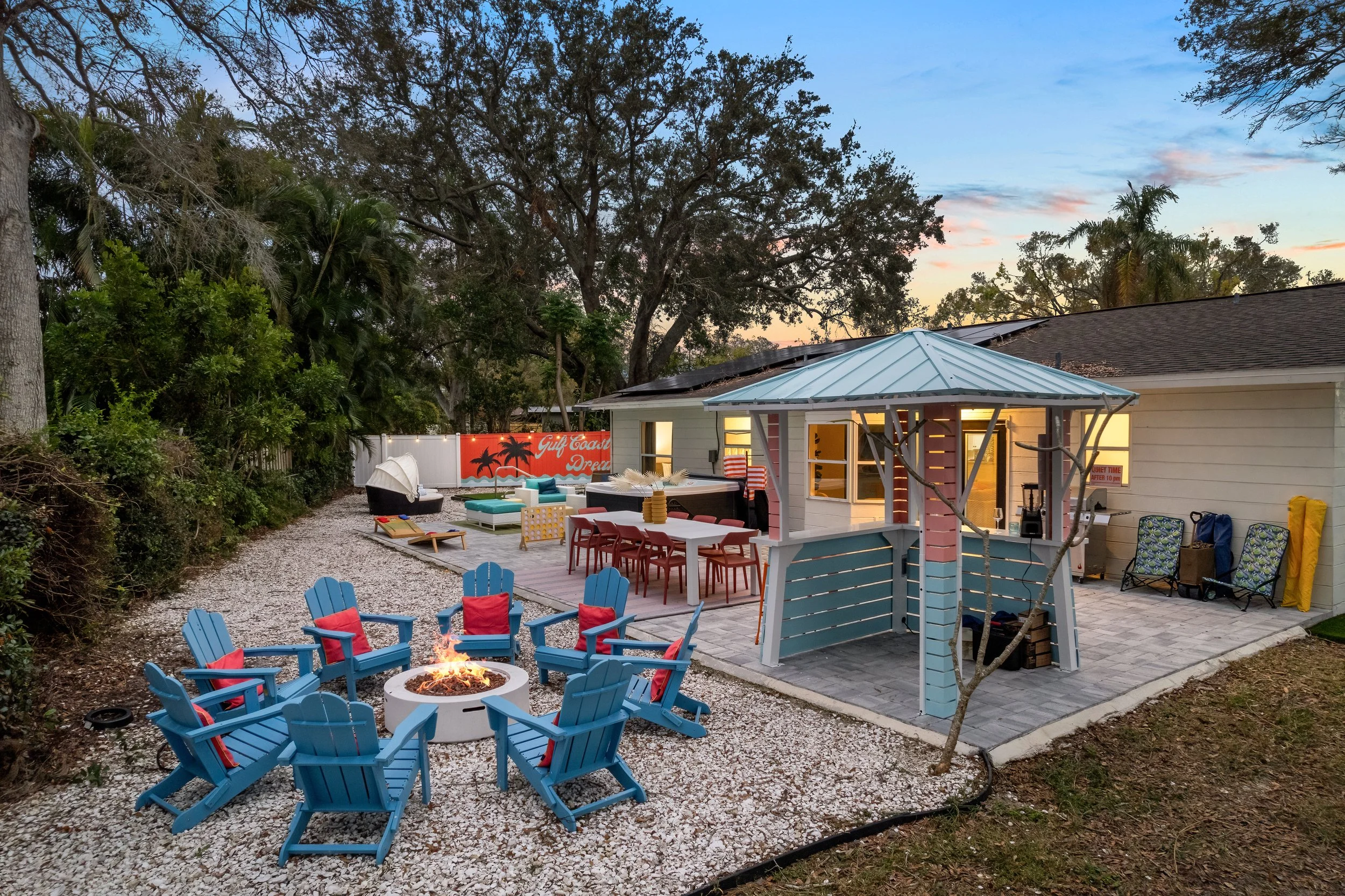 Backyard with outdoor seating area, fire pit, and small gazebo, illuminated at sunset with trees and a house in the background.