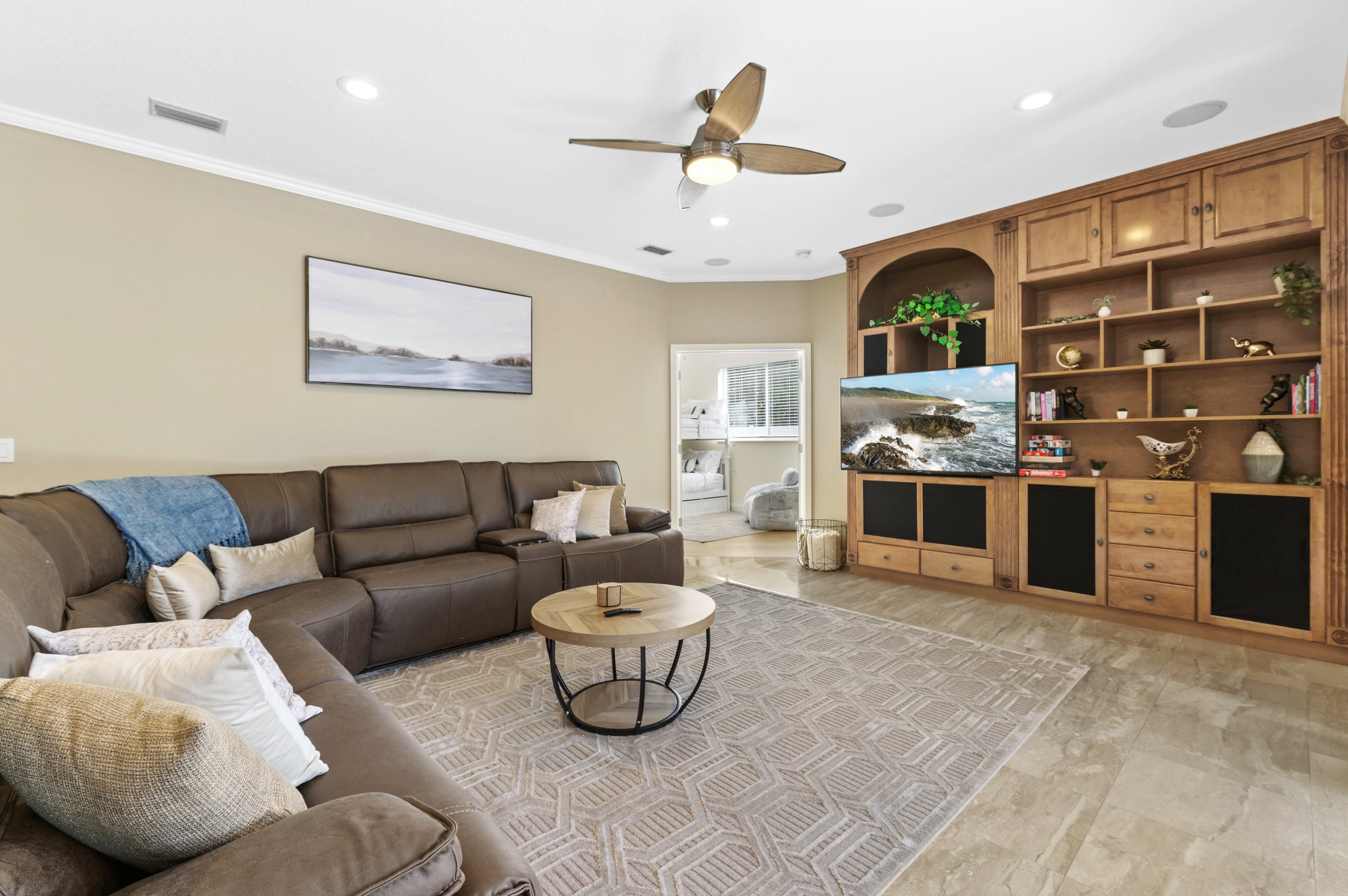 Living room with brown sectional couch, beige rug, round wooden coffee table, ceiling fan, built-in bookshelf and TV, decorative pillows, and coastal artwork on wall.