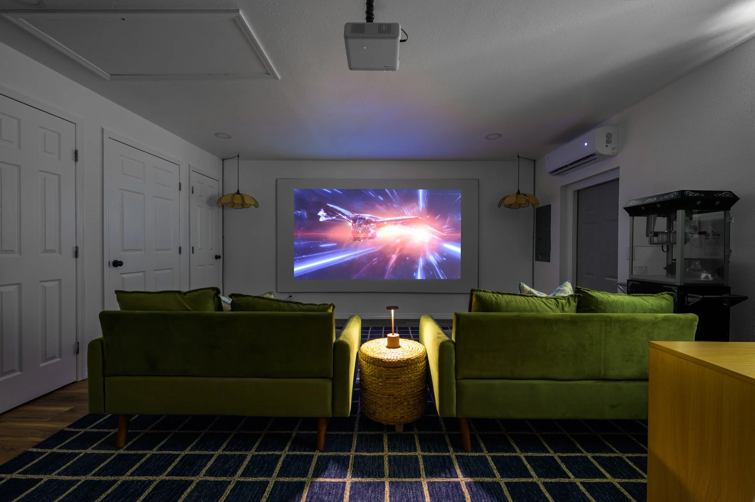 Home theater room with two green velvet sofas facing a projector screen displaying a space scene, a small woven side table with a lamp between the sofas, dark patterned carpet, white walls, and a popcorn machine to the right.