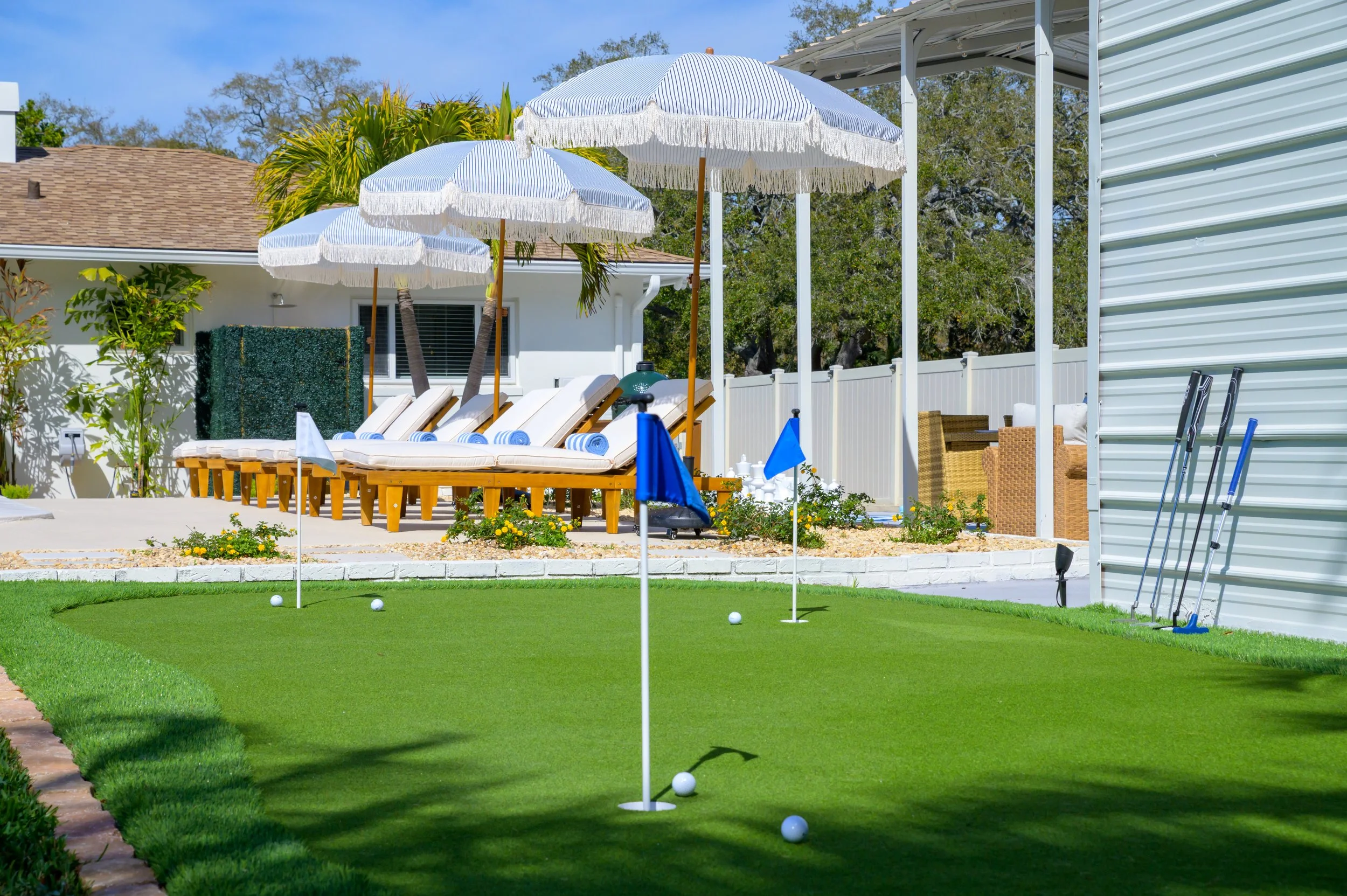 Backyard with a mini golf putting green, three golf balls, and a flag. In the background, there are four lounge chairs under striped umbrellas, a patio area with seating, and a white fence. There are trees and a house visible in the background.