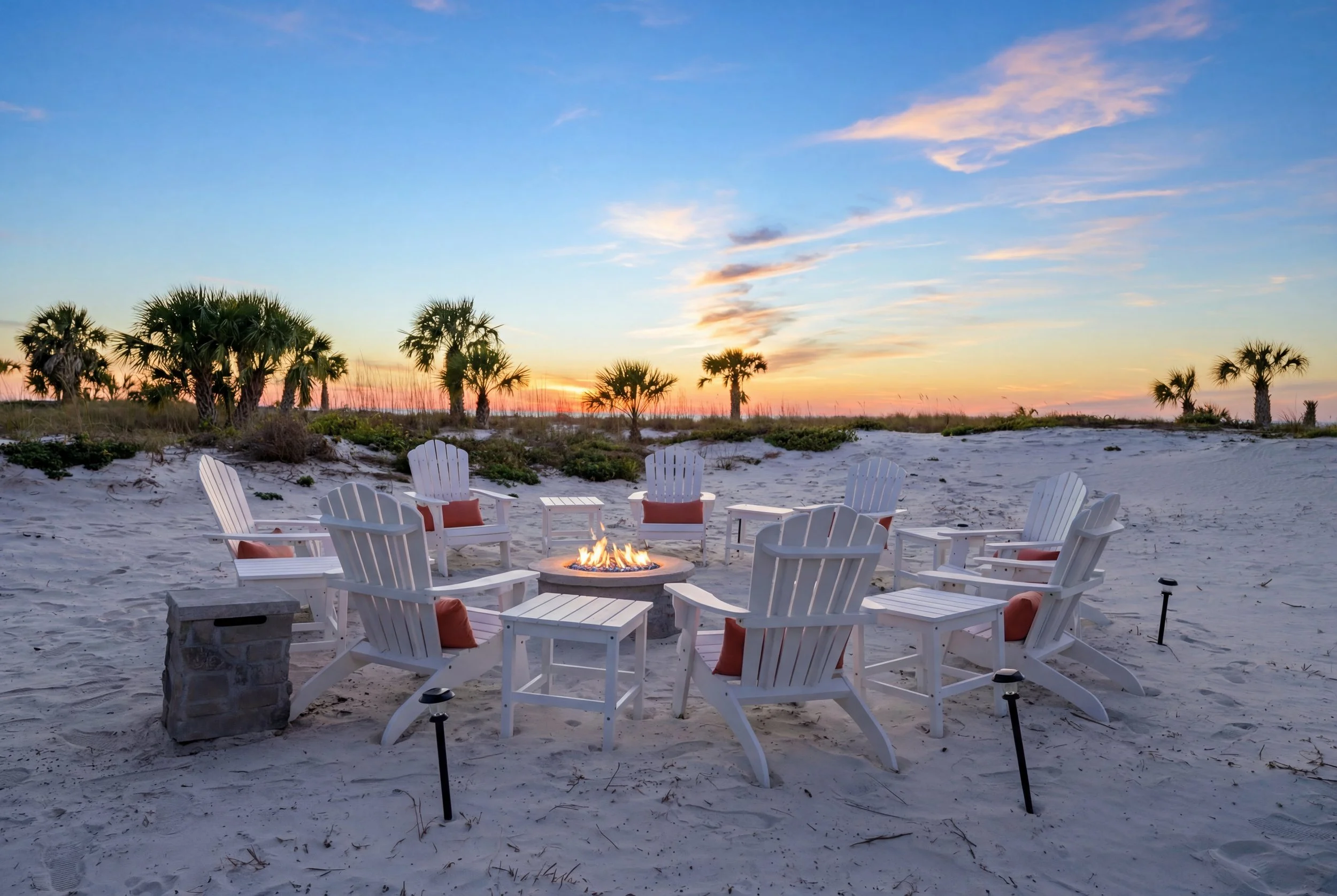 Beach with white Adirondack chairs arranged around a fire pit during sunset, with palm trees in the background.