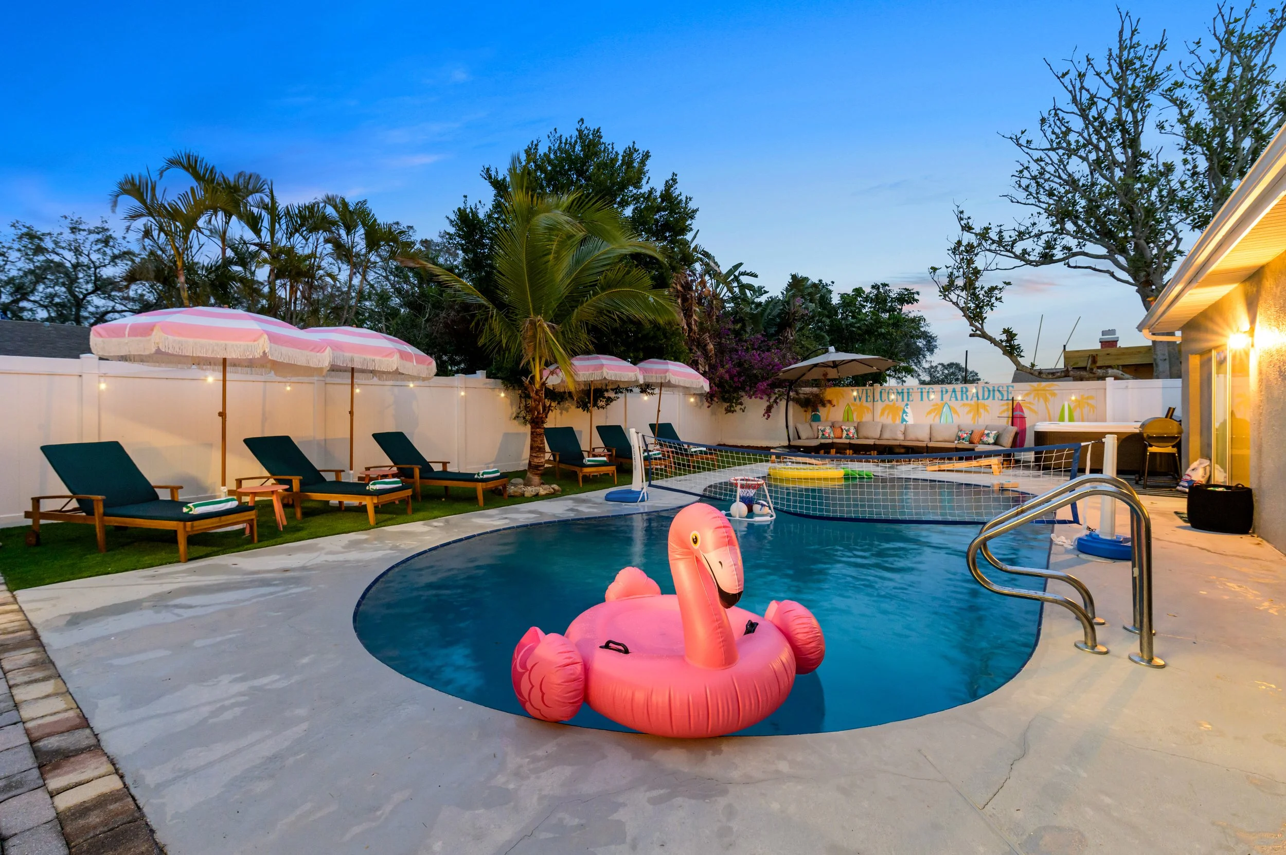 Backyard swimming pool with pink flamingo float, surrounded by lounge chairs with pink and white umbrellas, a net for volleyball, and outdoor seating under a sign that reads 'Welcome to Paradise' during dusk.