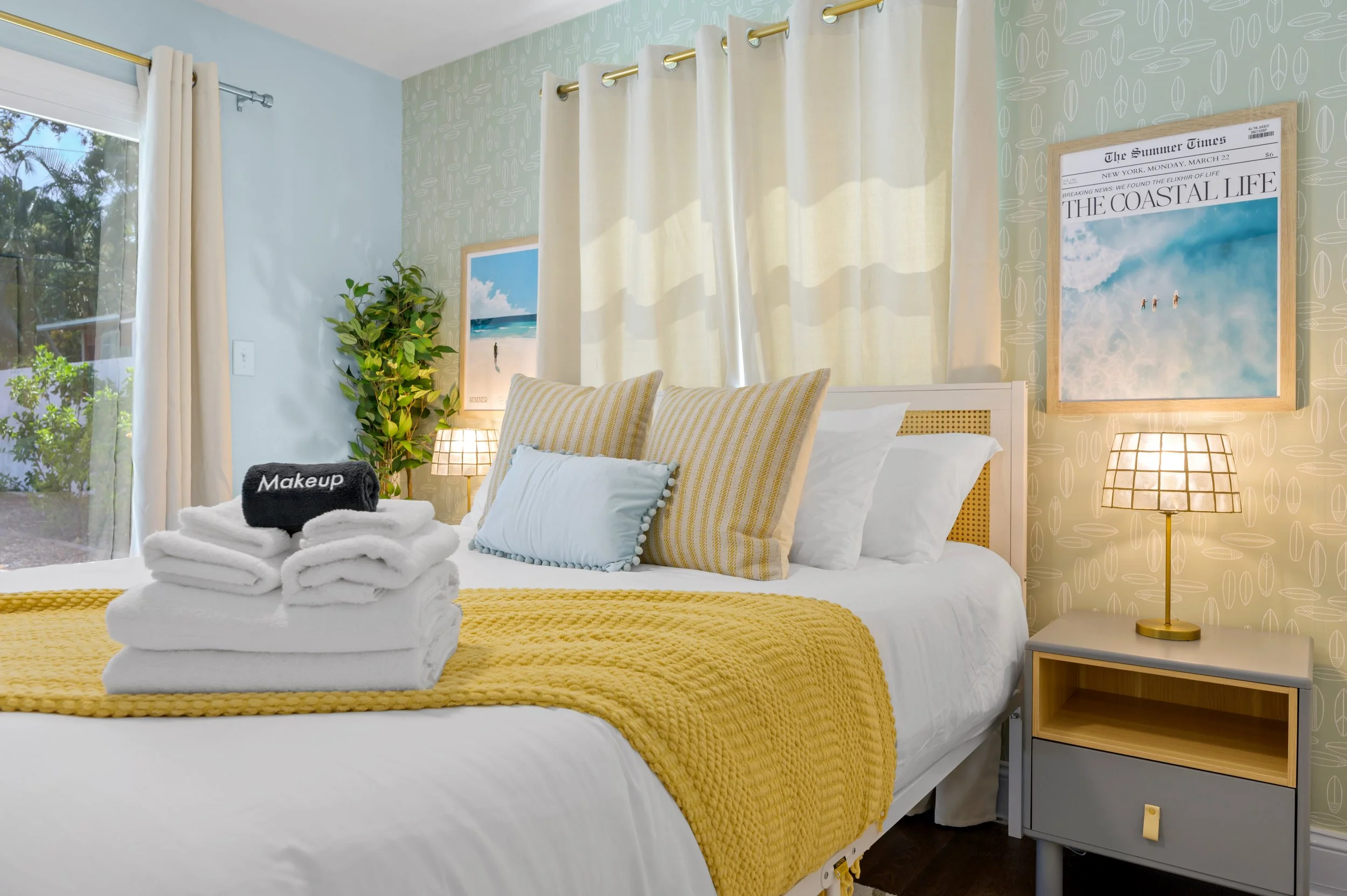 A neatly made bed with white linens, accented with yellow and light blue pillows, and a yellow knitted blanket. On the bed are folded white towels with a black roll labeled 'Makeup.' The room has a nightstand with a lamp, a large window with beige cu