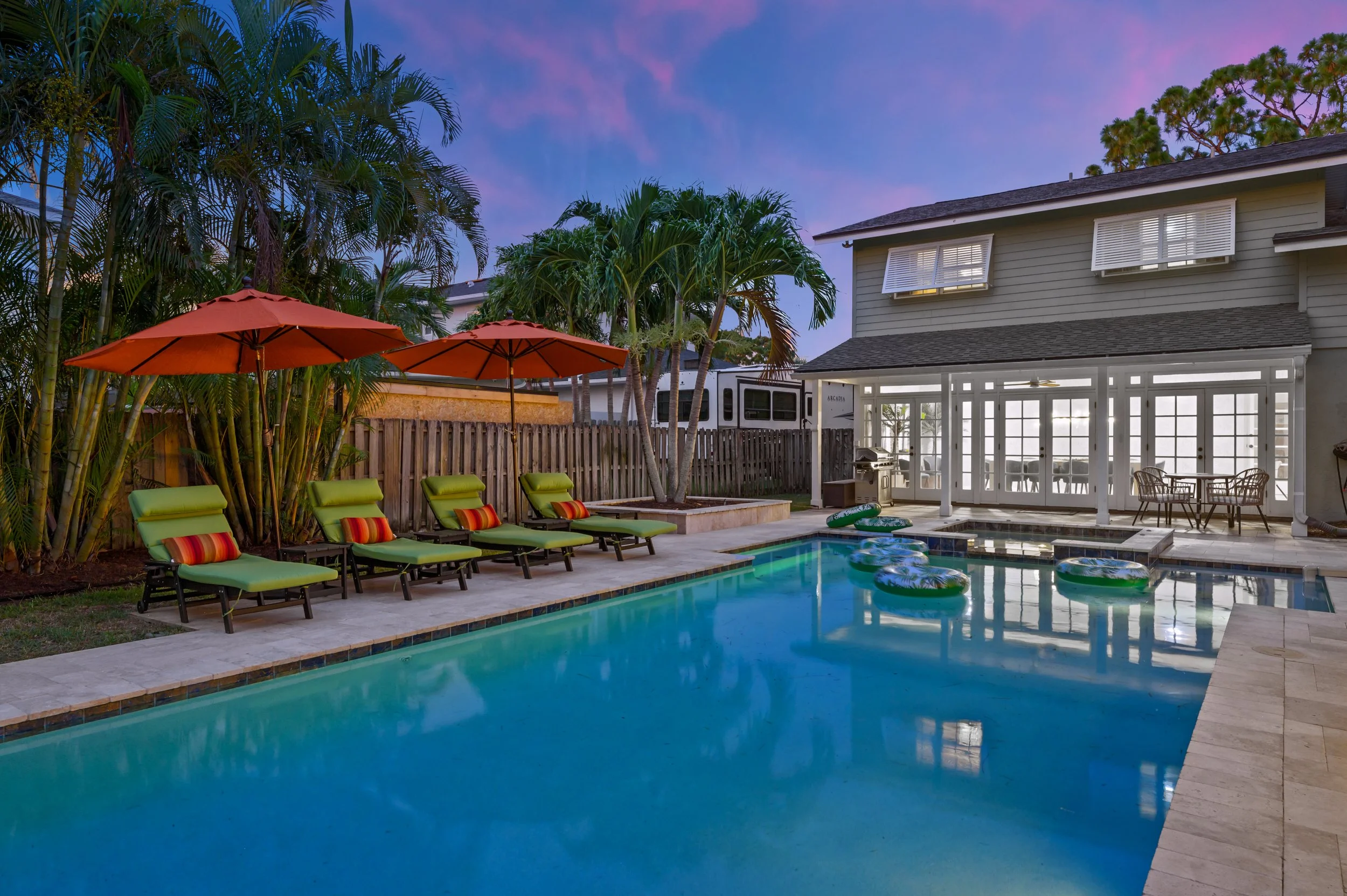 A backyard swimming pool area at dusk, with four green lounge chairs with striped pillows and orange umbrellas, surrounded by tropical plants and a house with a covered patio with glass doors.