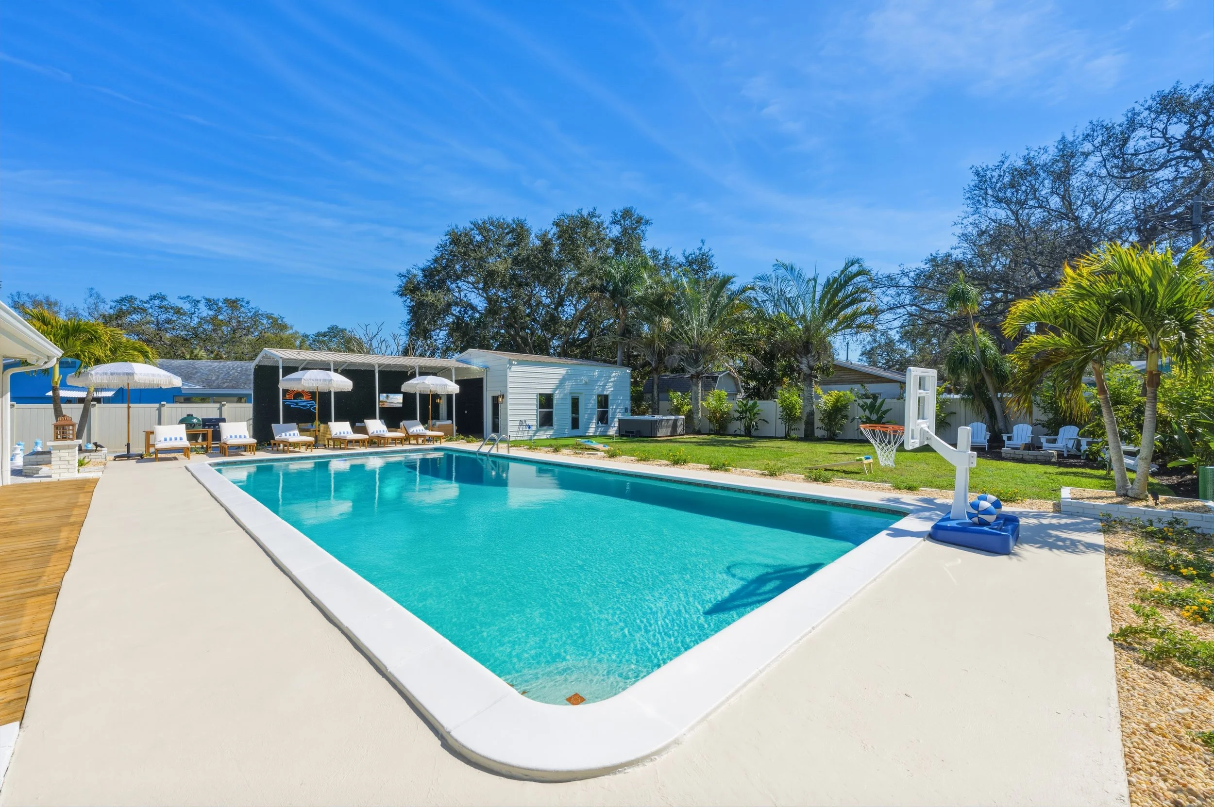 Backyard with a swimming pool, lounge chairs with umbrellas, palm trees, leafy bushes, and a basketball hoop, on a sunny day with a blue sky.