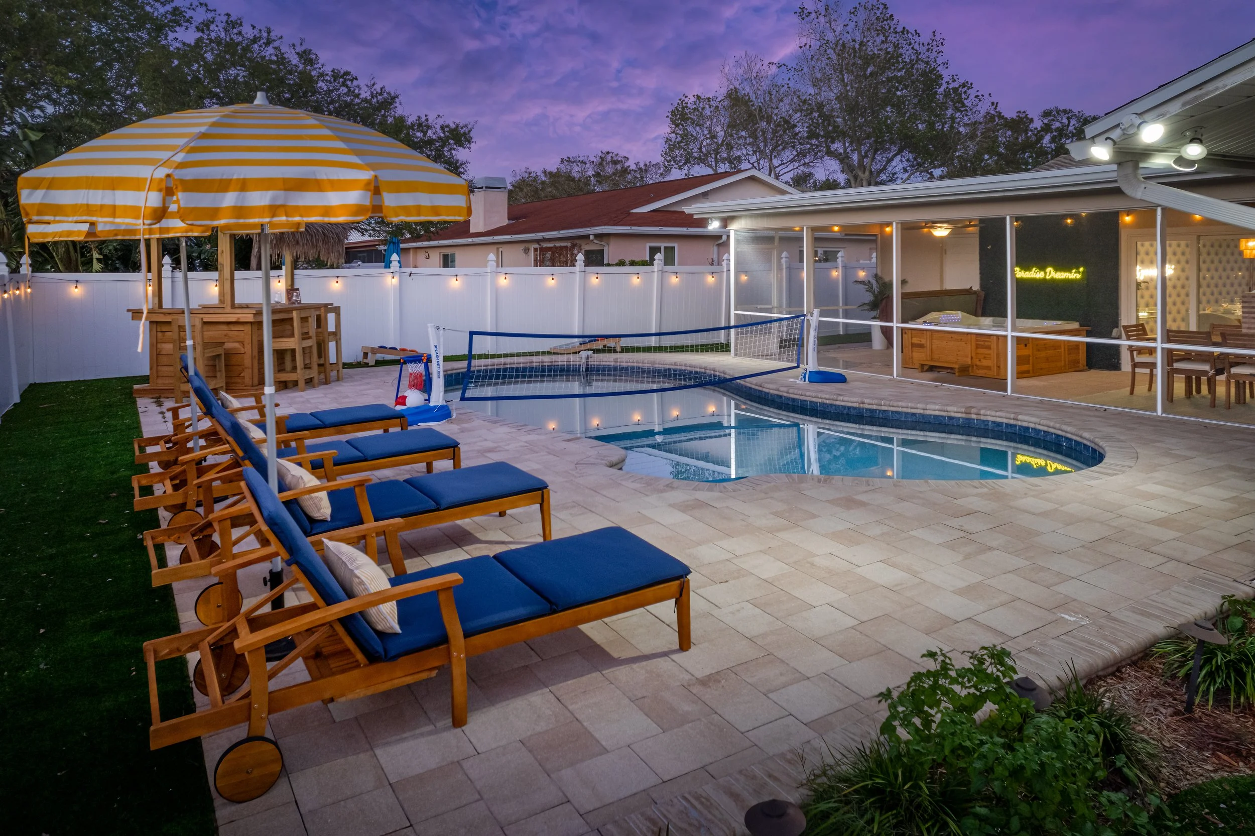 Backyard with swimming pool, lounge chairs with blue cushions, screened porch with seating, volleyball net, and string lights at dusk.
